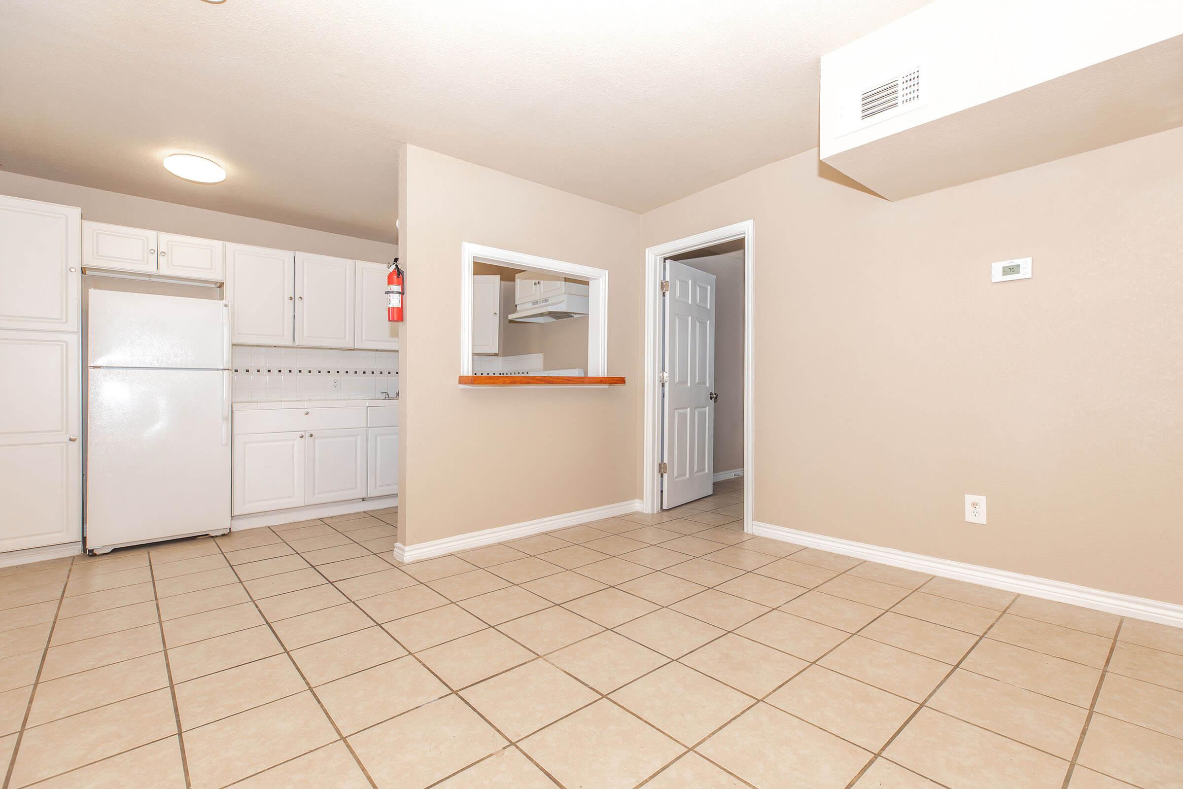 A spacious, well-lit kitchen area featuring white cabinets, a refrigerator, and a tile floor. A doorway leads to another room, and a small bar area opens to the kitchen. The walls are painted a light beige, creating a warm ambiance. A fire extinguisher is mounted on the wall for safety.