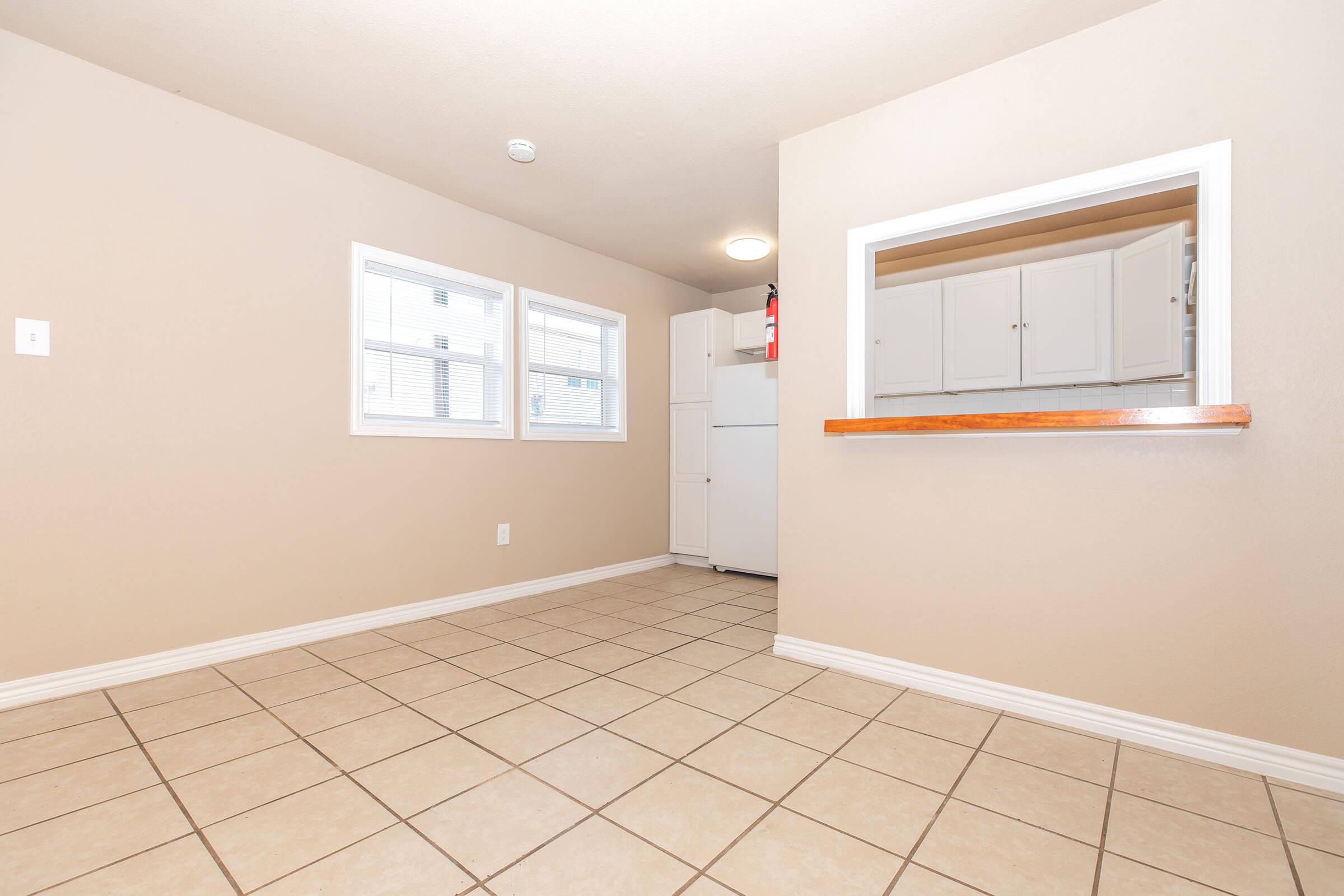 A light beige room with tiled flooring, featuring two windows on one wall. A small opening leads to a kitchen area with white cabinets and a refrigerator visible. The walls are plain, and the ceiling has a light fixture. The overall atmosphere is clean and simple, suitable for a dining or living space.