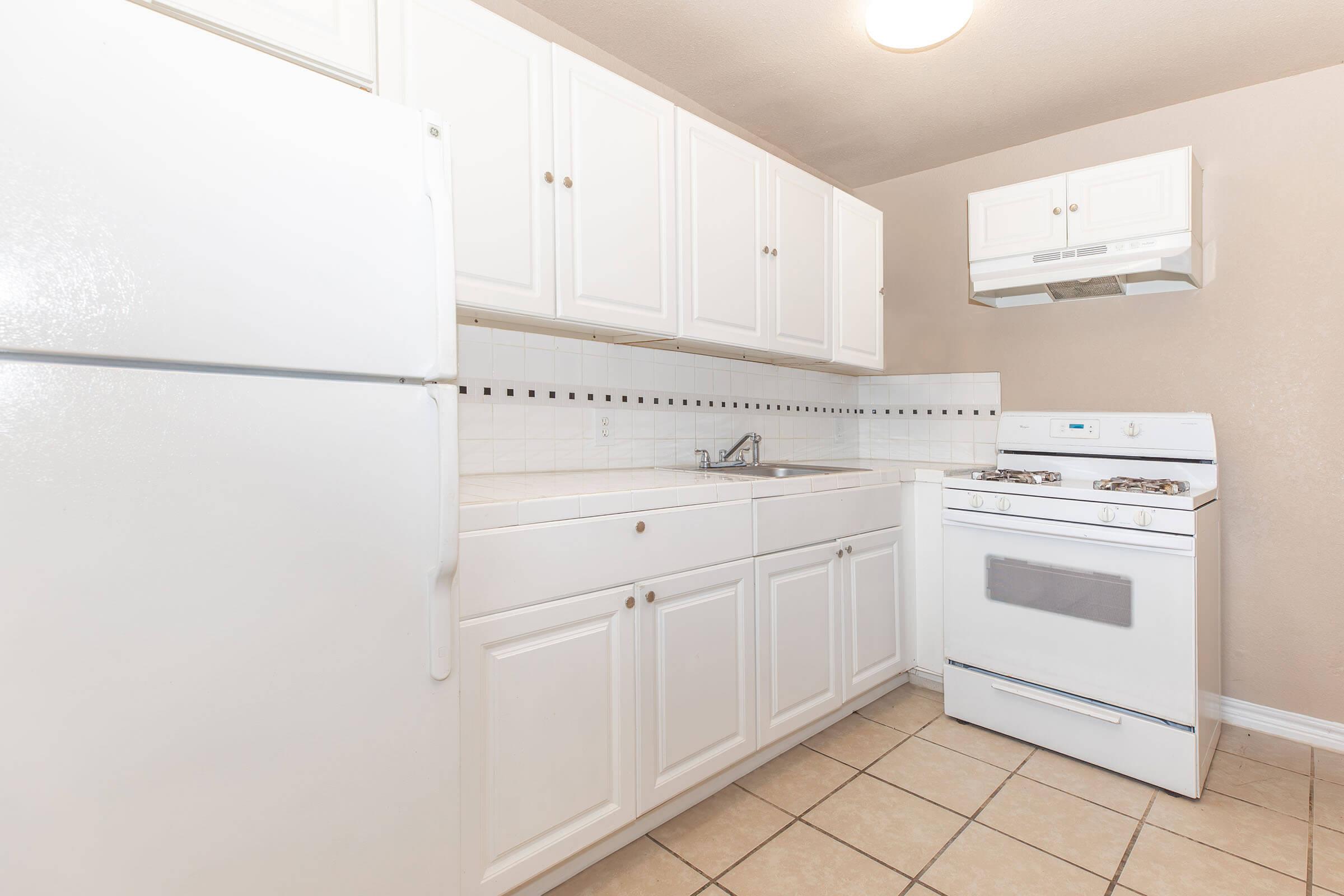 A small, tidy kitchen featuring white cabinetry, a double sink, and a white gas stove. The space includes a white refrigerator and beige tile flooring. A microwave is mounted above the stove, and natural light is provided by a ceiling fixture. The walls are painted a light beige, creating a clean and bright atmosphere.