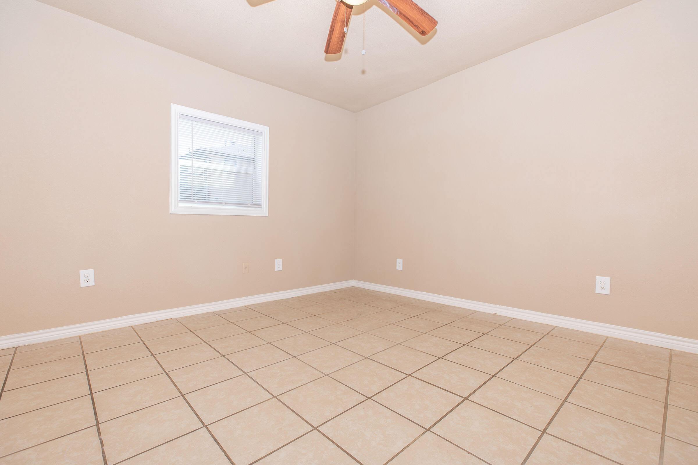 Empty room with beige walls and tile flooring. A single window with blinds on one wall allows natural light. The ceiling features a wooden ceiling fan. No furniture or decorations are present, emphasizing the spaciousness of the area.