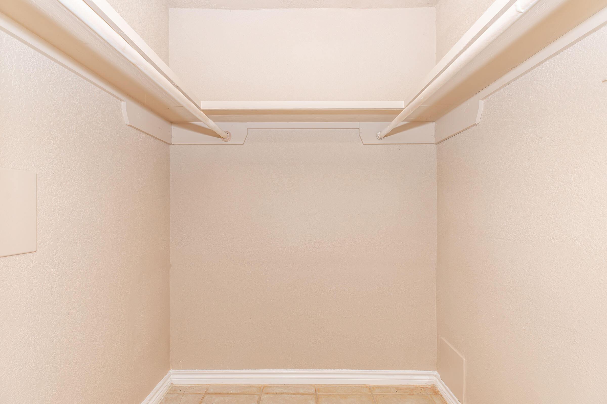 Empty closet with beige walls and a tiled floor. Two horizontal rods for hanging clothes are mounted on the walls, with no shelves or other storage items visible.