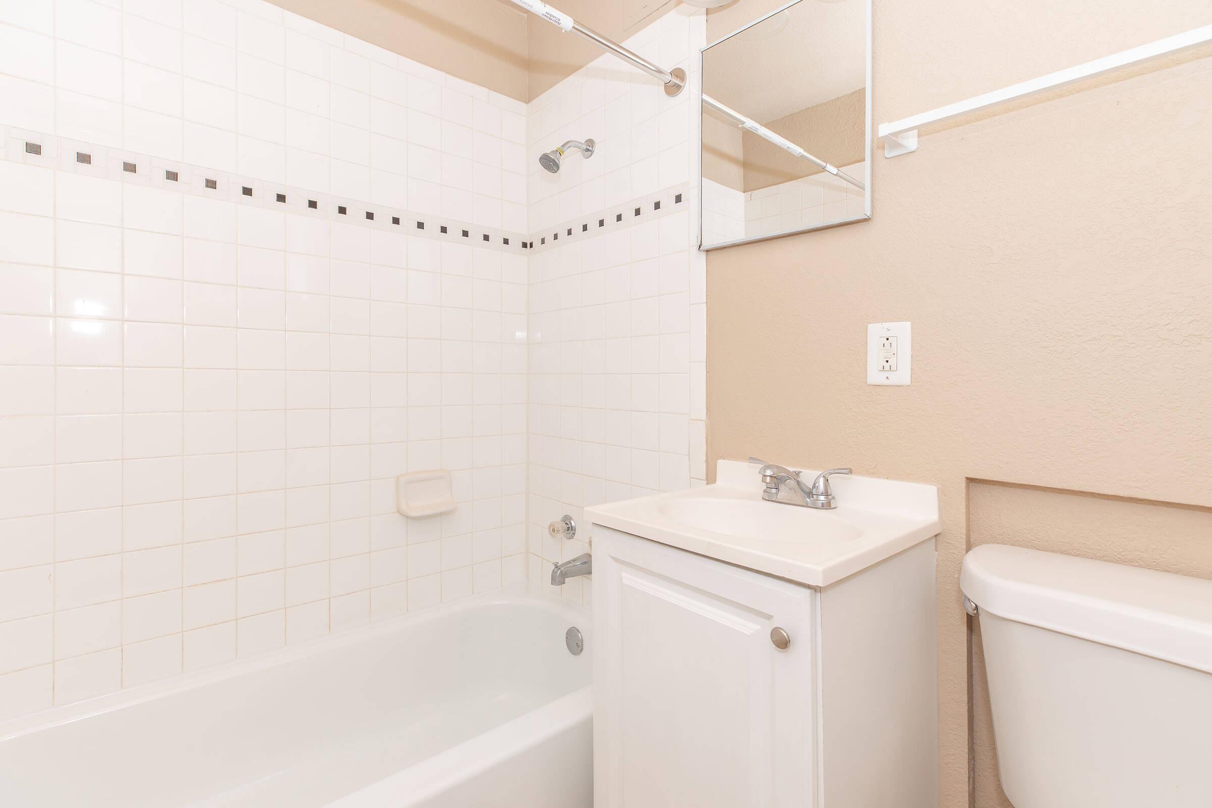 A clean and minimalistic bathroom featuring a white tiled shower and bathtub. A small white vanity with a sink is on the left, a mirror is mounted above it, and a toilet is visible on the right. The walls are painted a light beige color, creating a bright and airy atmosphere.