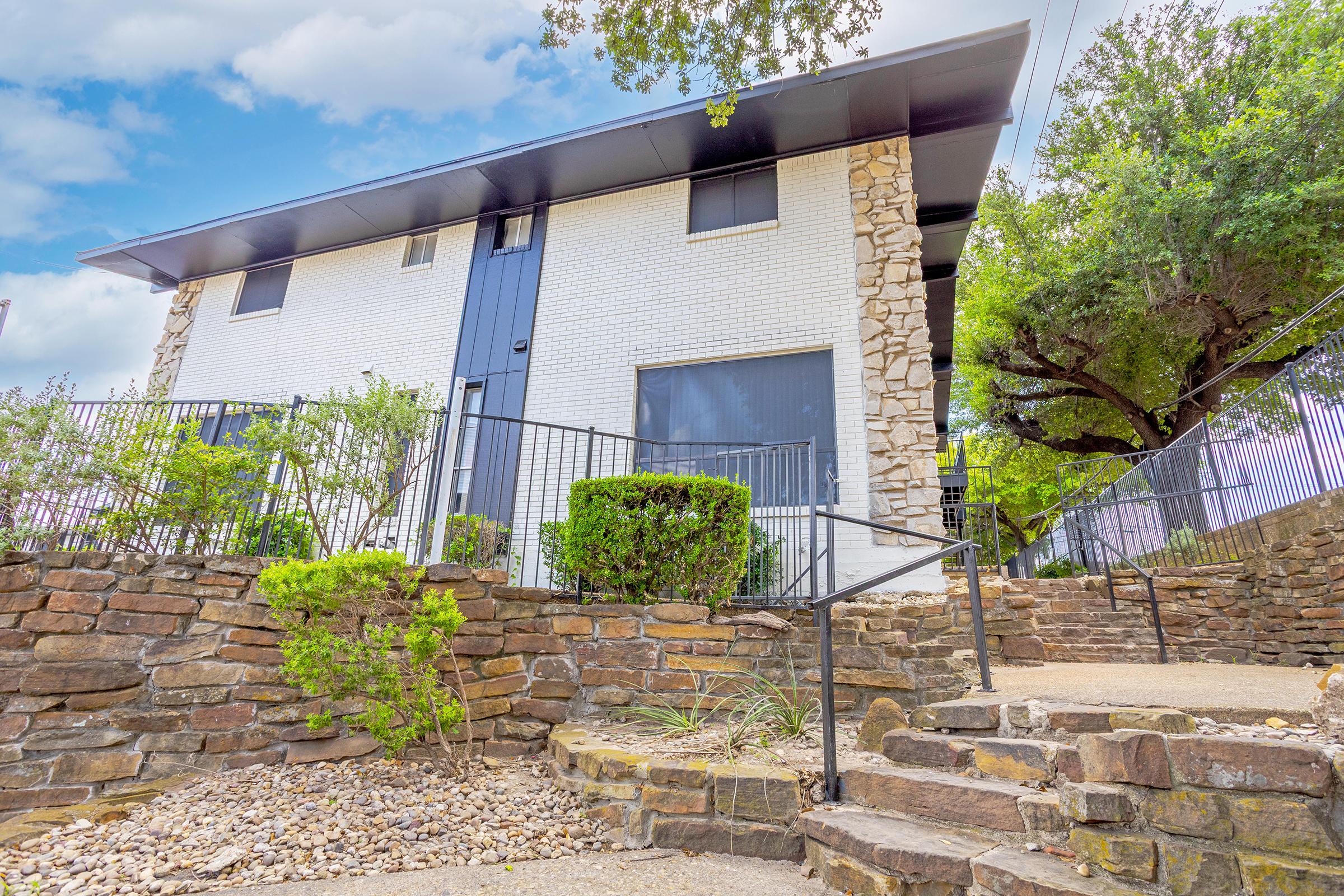 A modern two-story building with a stone exterior and large windows, surrounded by greenery and a stone retaining wall. A pathway with stairs leads up to the entrance, with a wrought iron railing. The sky is partly cloudy, enhancing the overall brightness of the scene.