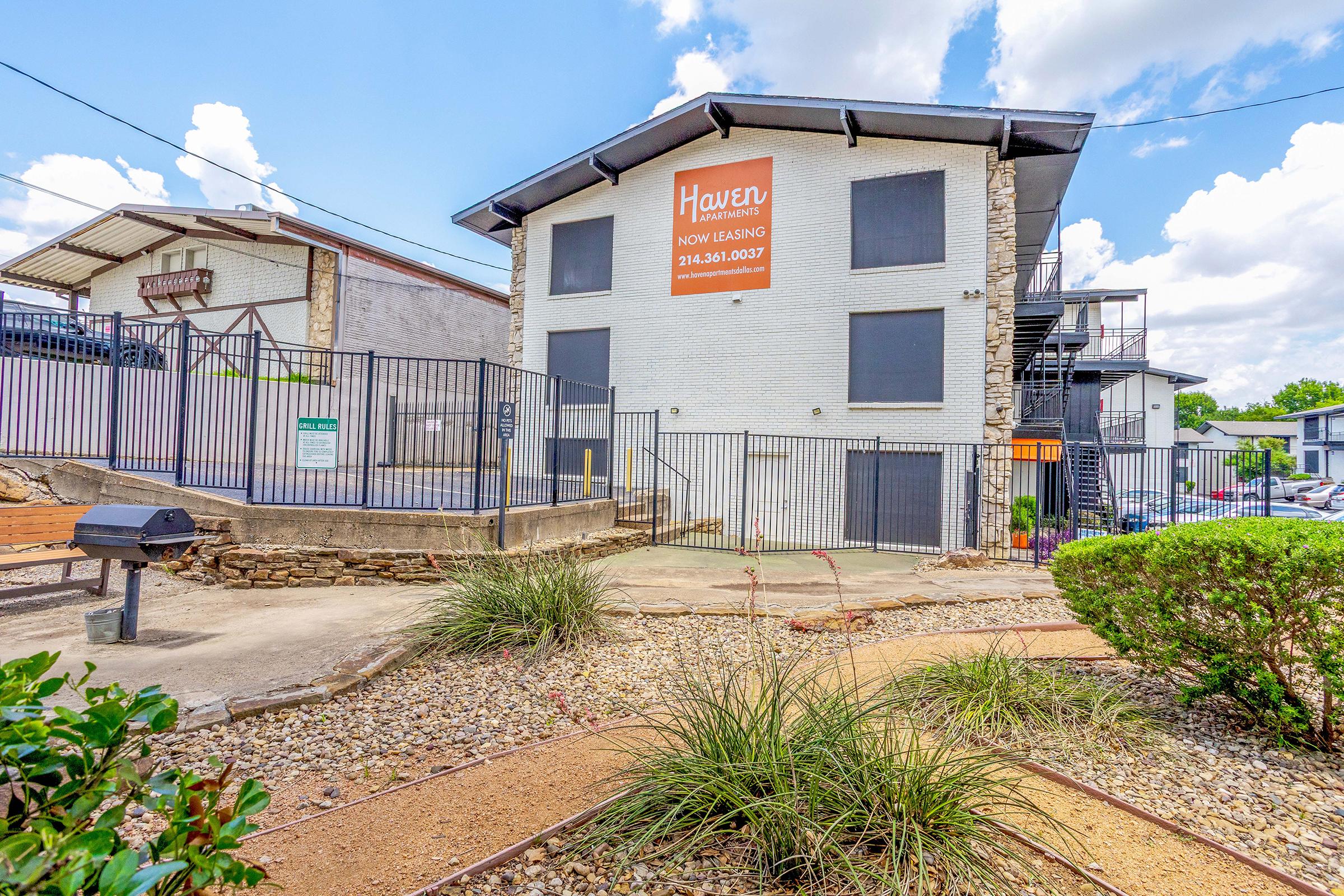 A modern apartment building with a white exterior and a prominent "Haven Now Leasing" sign. The property features a fenced area, landscaped pathways, and a grilling station. Nearby, there are multiple stories of the building visible, along with cloudy skies above.