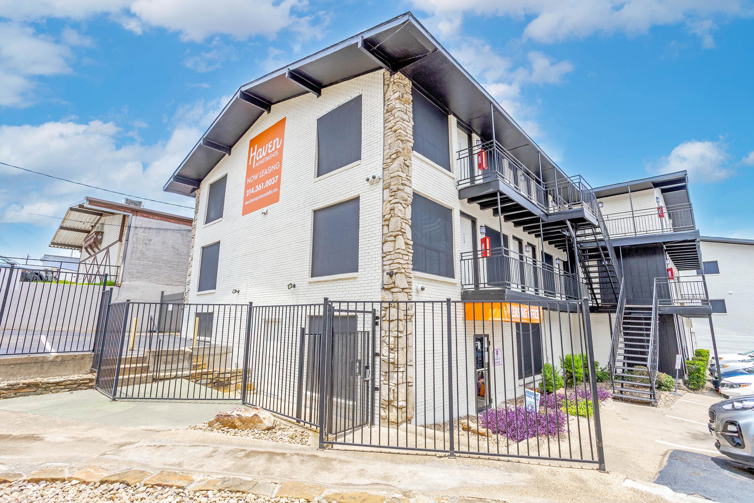 A modern multi-story building with a stone accent and large windows, featuring a gated entrance and vibrant purple flowers in the foreground. The building displays an orange sign with the word "Haven" prominently. A staircase leads to the upper levels, and parked cars are visible nearby under a bright blue sky.
