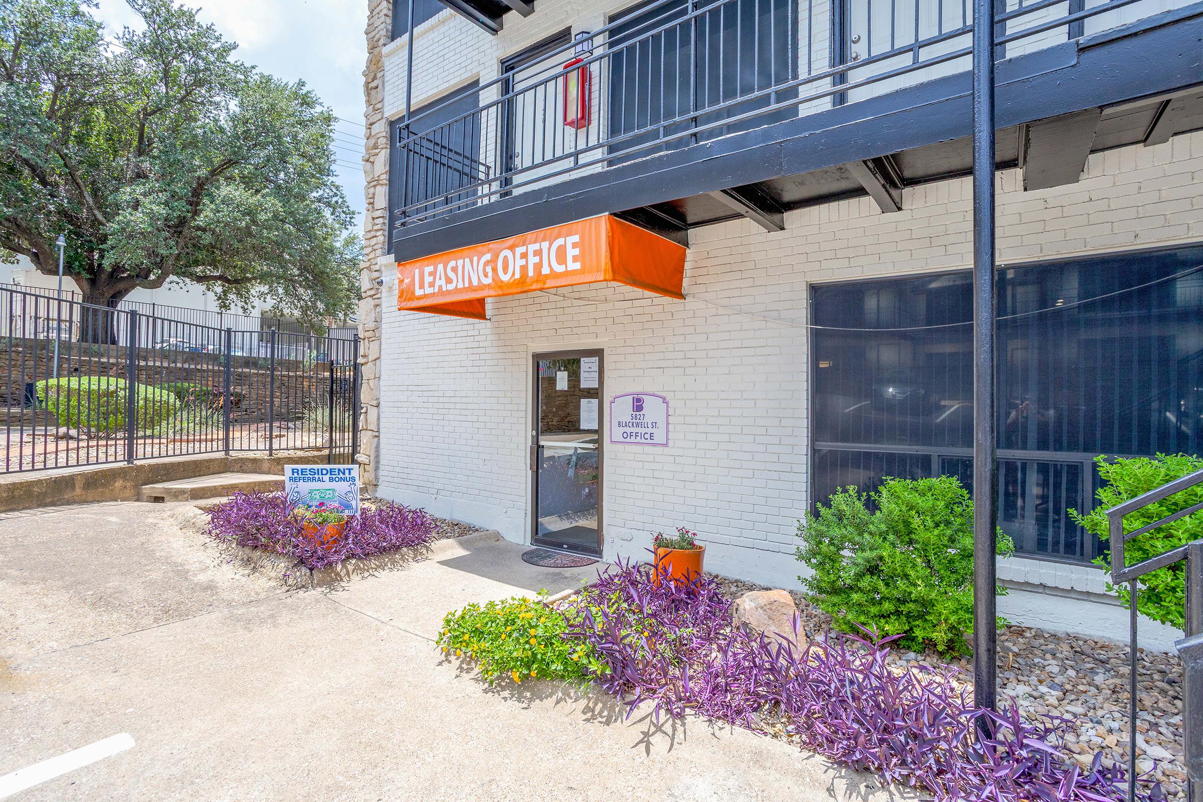 Leasing office entrance with a bright orange awning. The door features a sign indicating "Leasing Office." Surrounding greenery includes purple and green plants, with a sign in front that reads "Resident" near a sidewalk. The building has a white brick exterior and a gated area behind it.