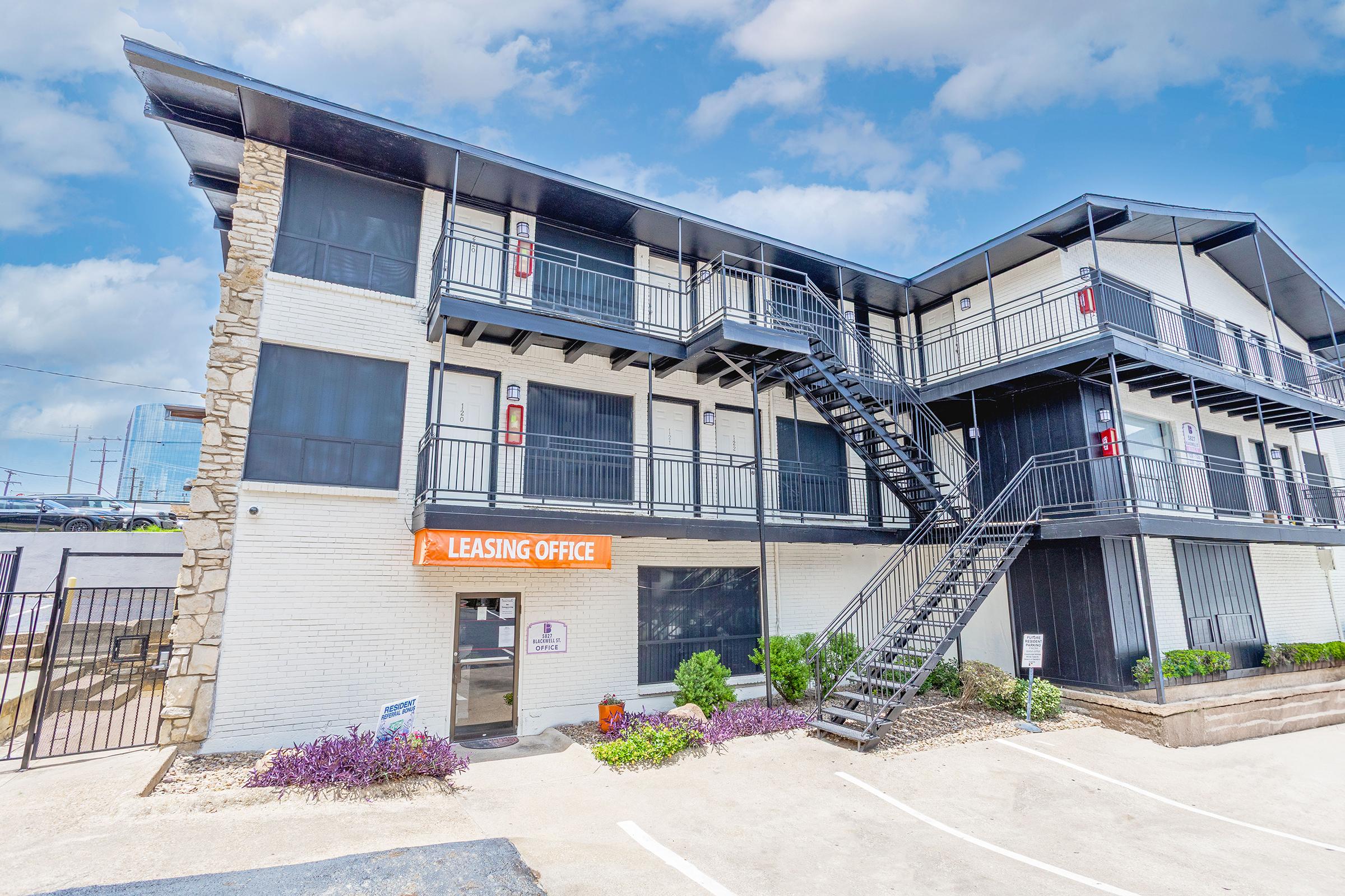 A three-story building featuring a leasing office with an orange sign. Staircases on the exterior lead to upper floors. Surrounded by landscaped areas with small plants and gravel. The building has a modern design with a mix of brick and metal elements, set against a partly cloudy sky.