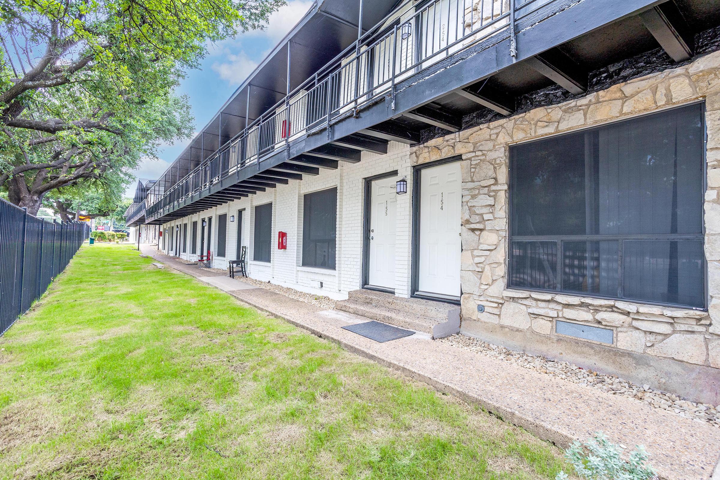A view of a multi-unit apartment building with a row of doors and balconies on the second floor. The exterior features a combination of stone and white siding, with a green lawn area in front. Trees are visible in the background, and the setting appears well-maintained and inviting.
