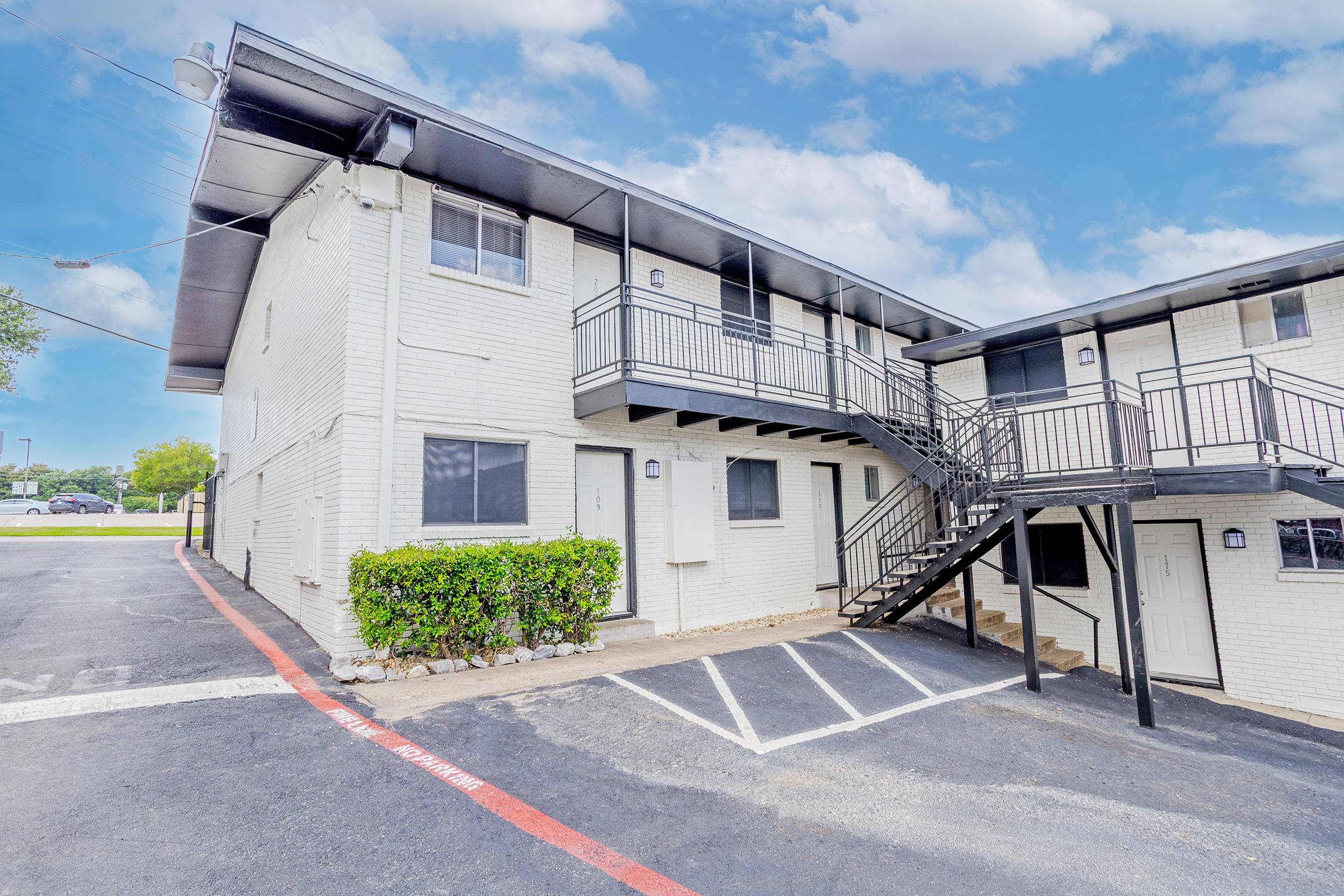 A two-story apartment building featuring white brick exterior, black railings, and a staircase. The image shows a paved parking area with marked spaces and greenery near the building. The sky is partly cloudy, adding a bright atmosphere to the setting.