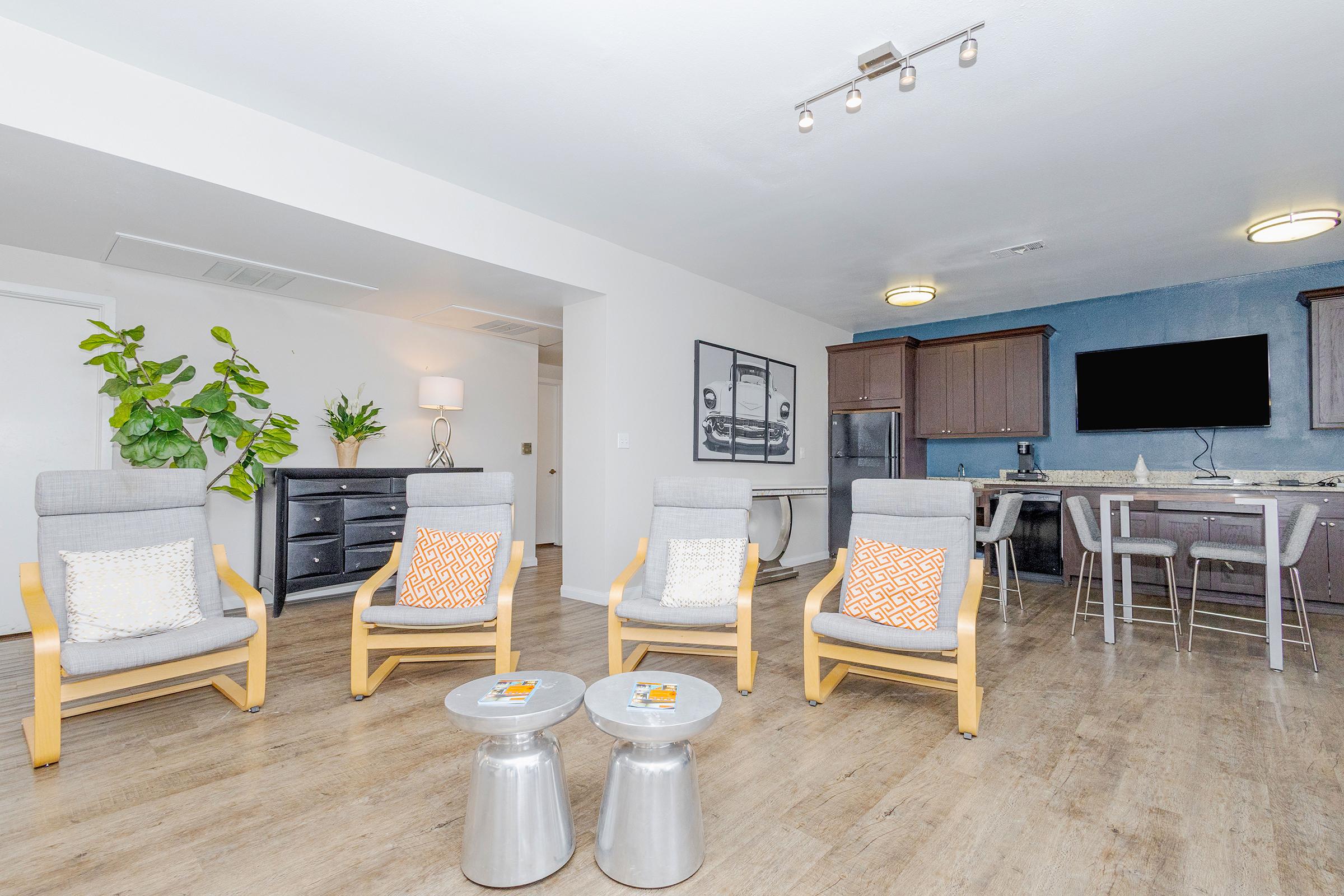 A modern living space featuring four light-colored lounge chairs with decorative pillows arranged in a circle. In the background, there is a sleek kitchen area with dark cabinetry, a television mounted on the wall, and a dining table. Green plants add a touch of nature to the room.