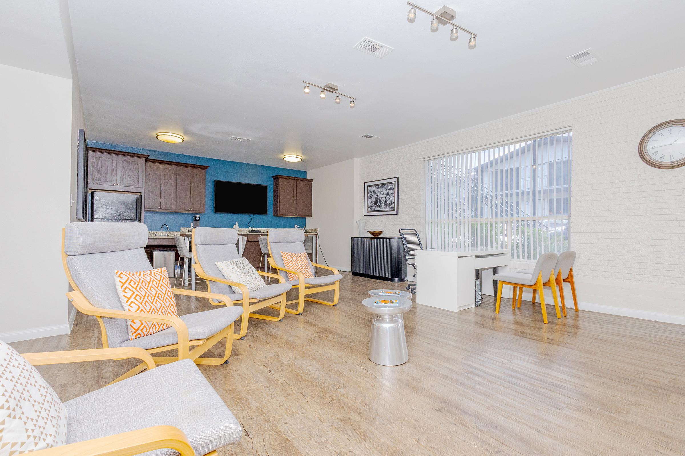 Bright and spacious common area featuring modern seating with light-colored wooden frames and gray cushions. There are kitchen facilities in the background, with a dining area. Natural light streams in through large windows, and decorative elements add a welcoming touch. A wall clock and artwork enhance the ambiance.