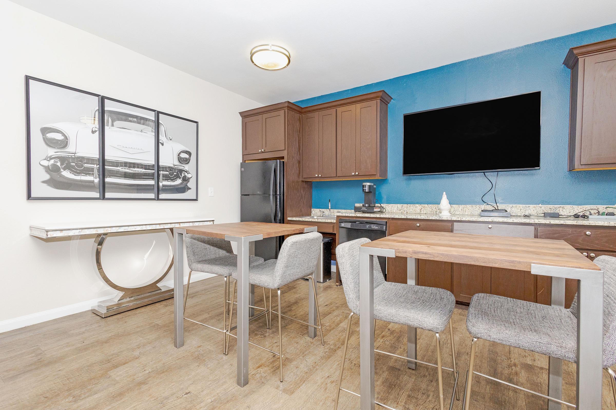 Modern kitchen featuring wooden cabinets, a large flat-screen TV mounted on a blue wall, and a sleek table with four stools. There is a stylish artwork of a vintage car on the wall, and the space has a contemporary feel with natural wood flooring.