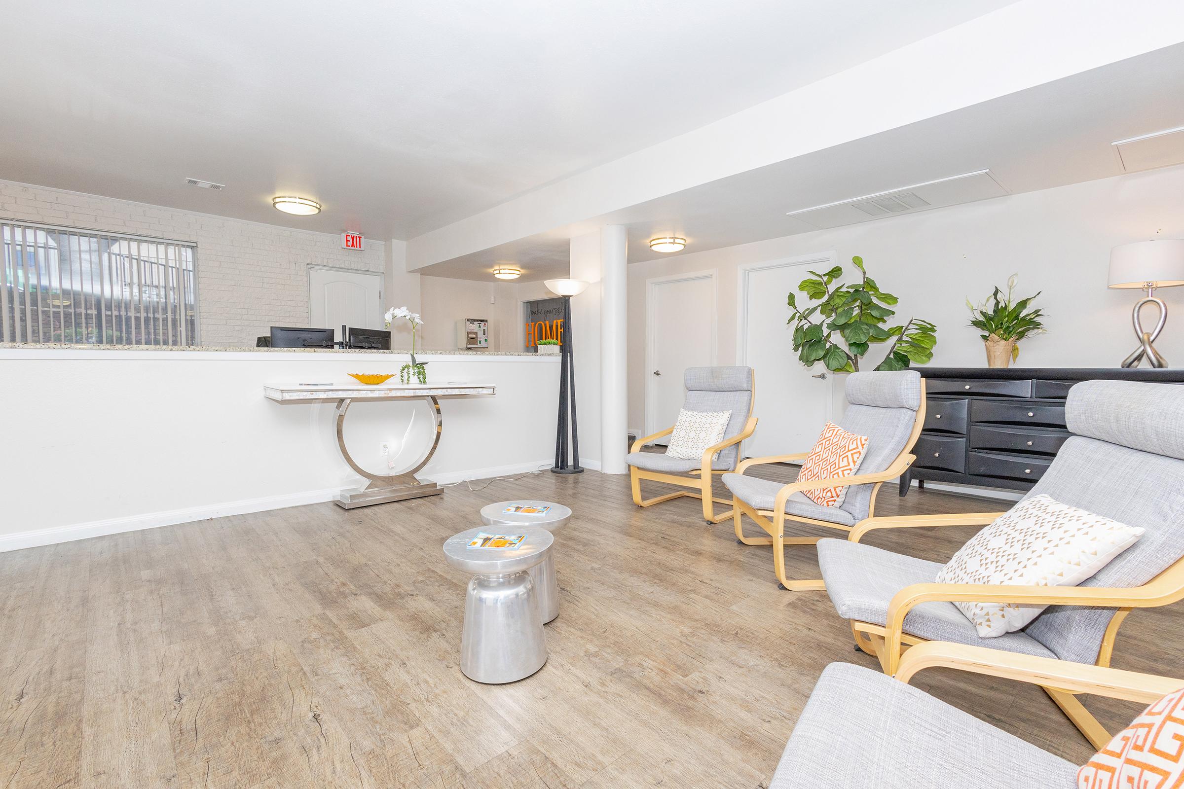 A modern waiting area featuring three wooden lounge chairs with cushions, two small metallic tables, and a reception desk in the background. The space has light-colored walls, wooden flooring, and decorative plants, creating a welcoming and comfortable atmosphere.
