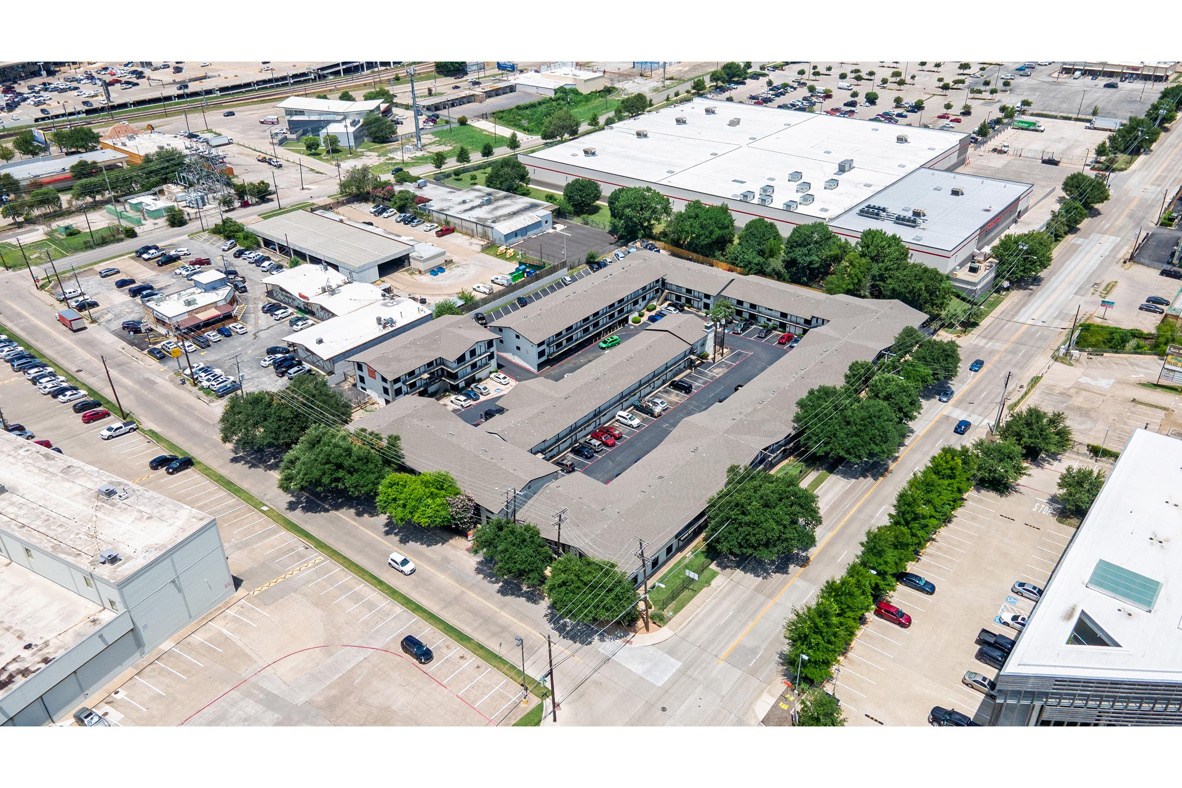 Aerial view of a large commercial complex with multiple buildings surrounded by a parking lot. Nearby, there are several other buildings and businesses, along with trees lining the streets. The image captures a bustling urban environment with a mix of vehicles and greenery.