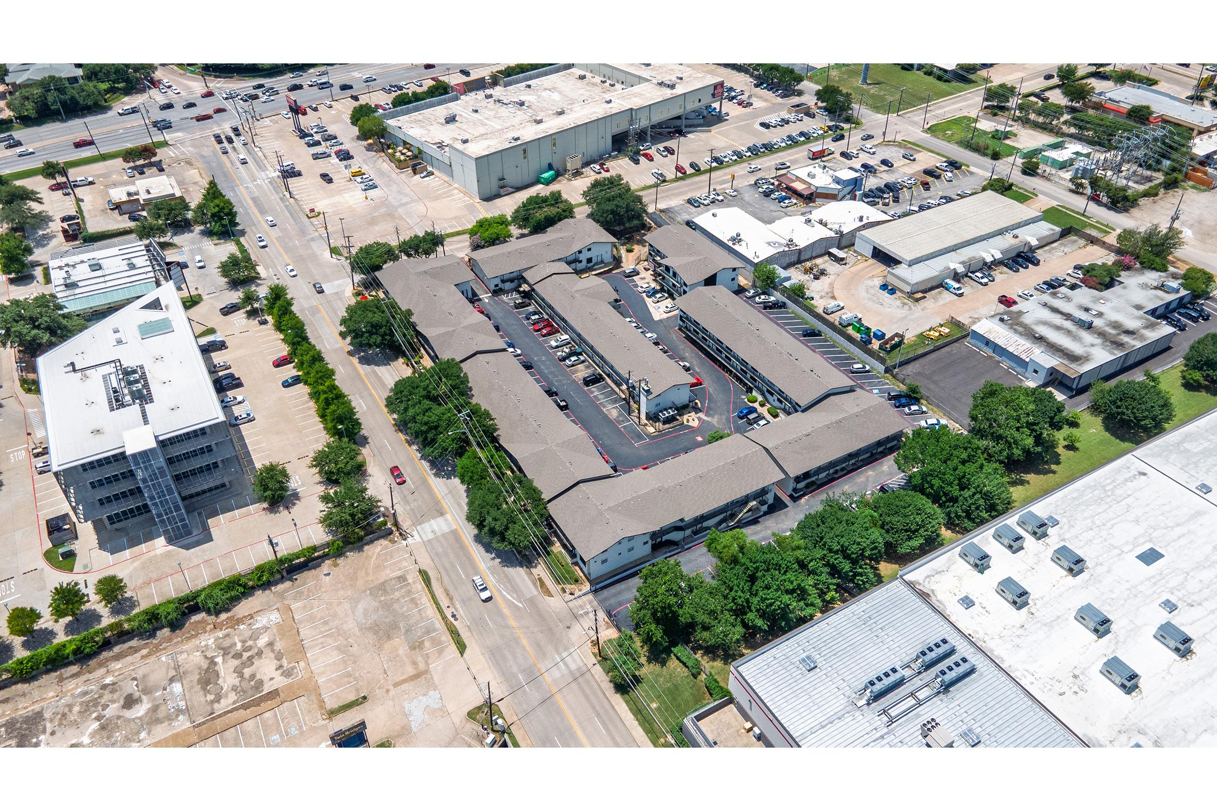 Aerial view of a commercial area featuring multiple buildings, parking lots, and roads. The image includes a mix of low-rise structures, green trees, and vehicles. Nearby, there are industrial buildings and a parking lot, showcasing a busy urban landscape.