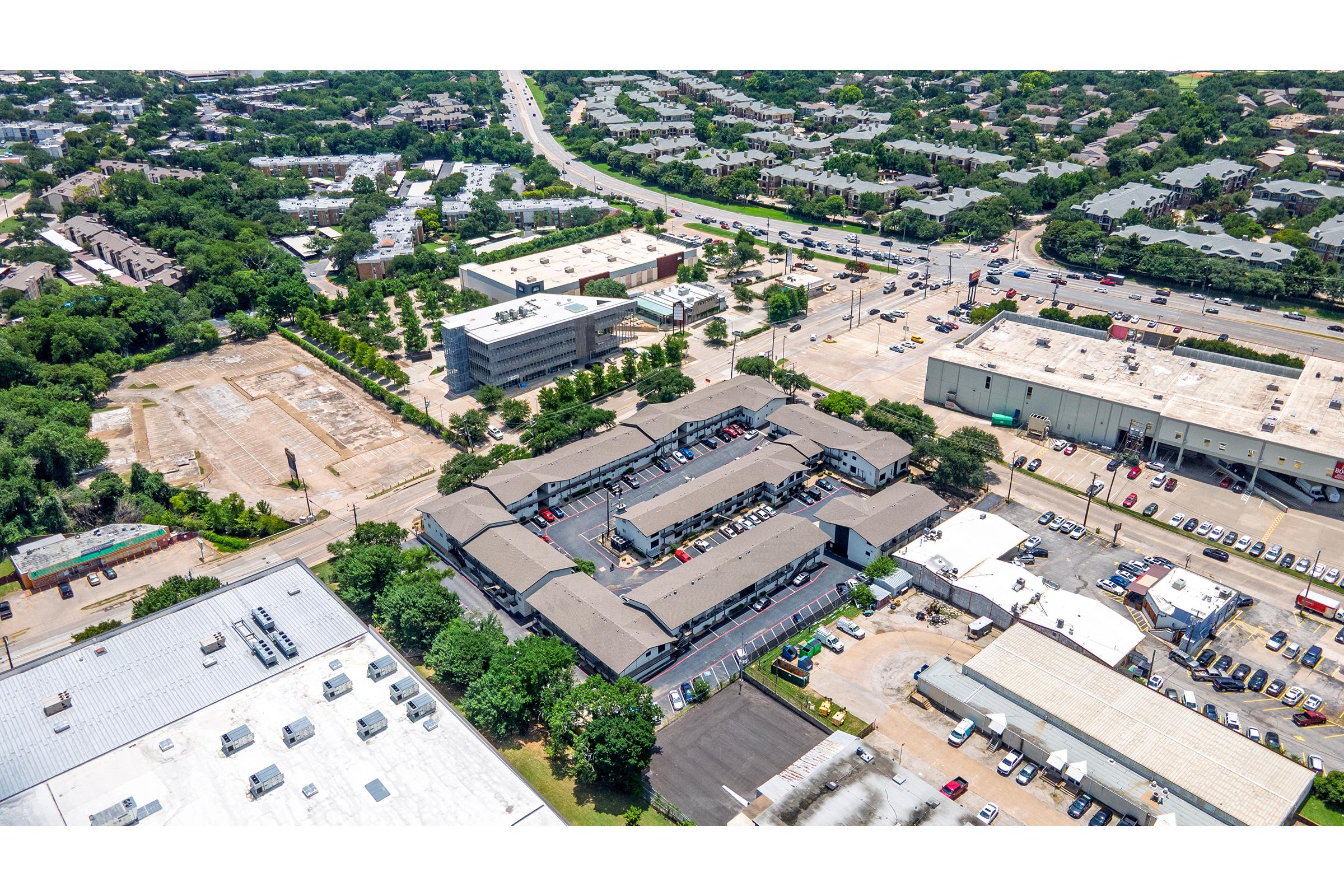 Aerial view of a commercial area featuring several buildings, parking lots, and surrounding greenery. In the foreground, a multi-unit structure is visible with parked vehicles. The background shows a mix of residential and commercial properties along a road, with open land and trees in between.