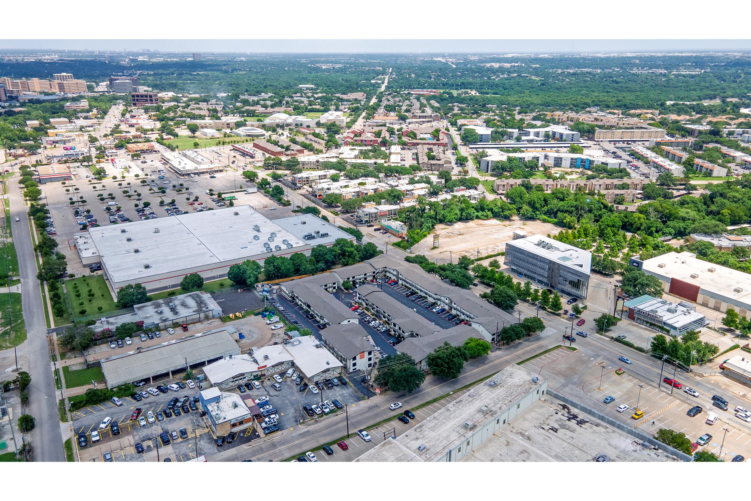 Aerial view of a large urban area featuring a mix of commercial and residential buildings, parking lots, and green spaces. The landscape shows a blend of developed areas and trees, with a skyline and roads visible in the distance, illustrating a vibrant city environment.