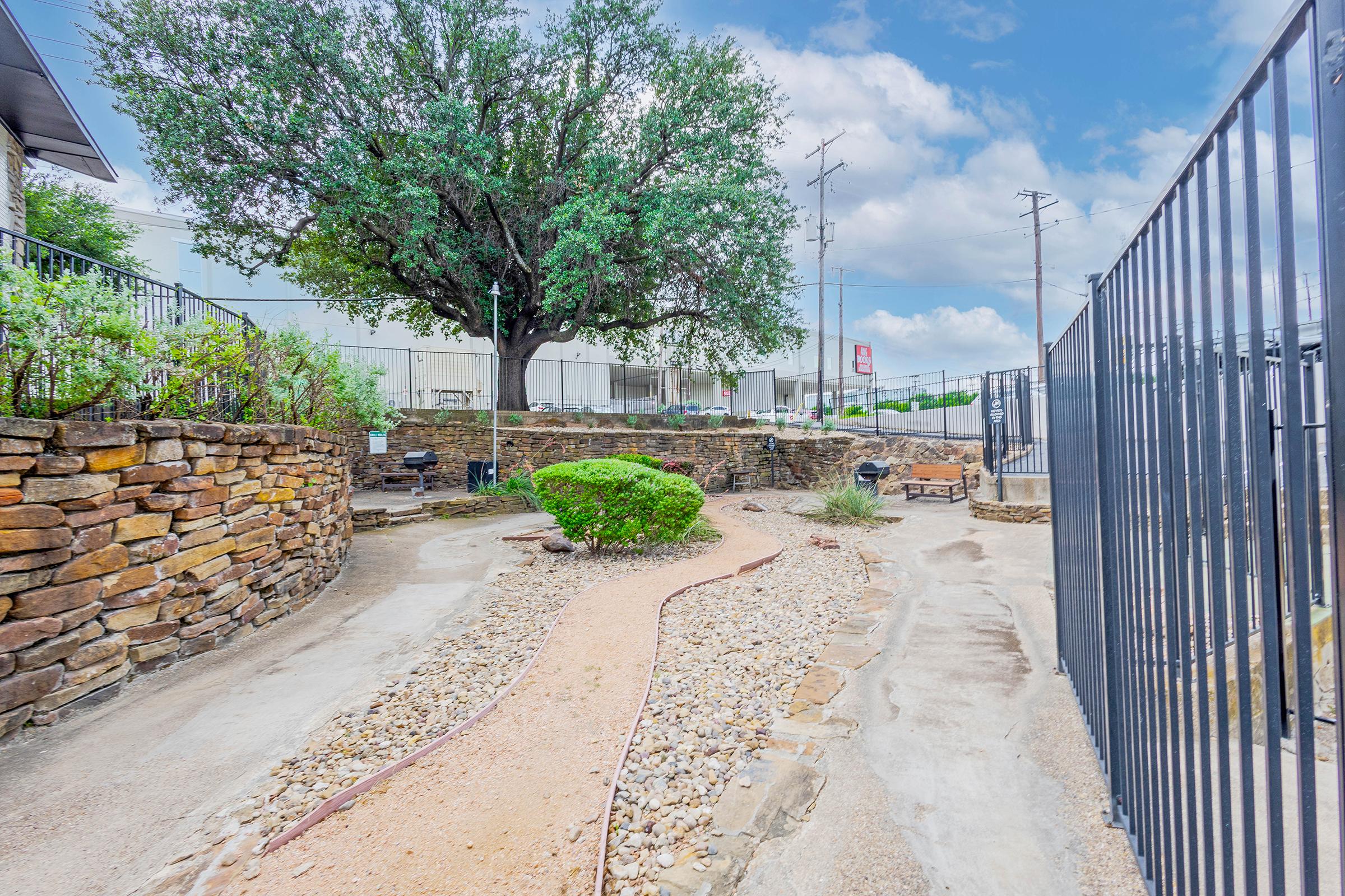 A peaceful outdoor pathway winding through a landscaped area with a large tree, stone borders, and low shrubs. The sky is partly cloudy, and there are utility poles in the background, suggesting an urban setting. A sense of tranquility and natural beauty is present in this garden-like space.