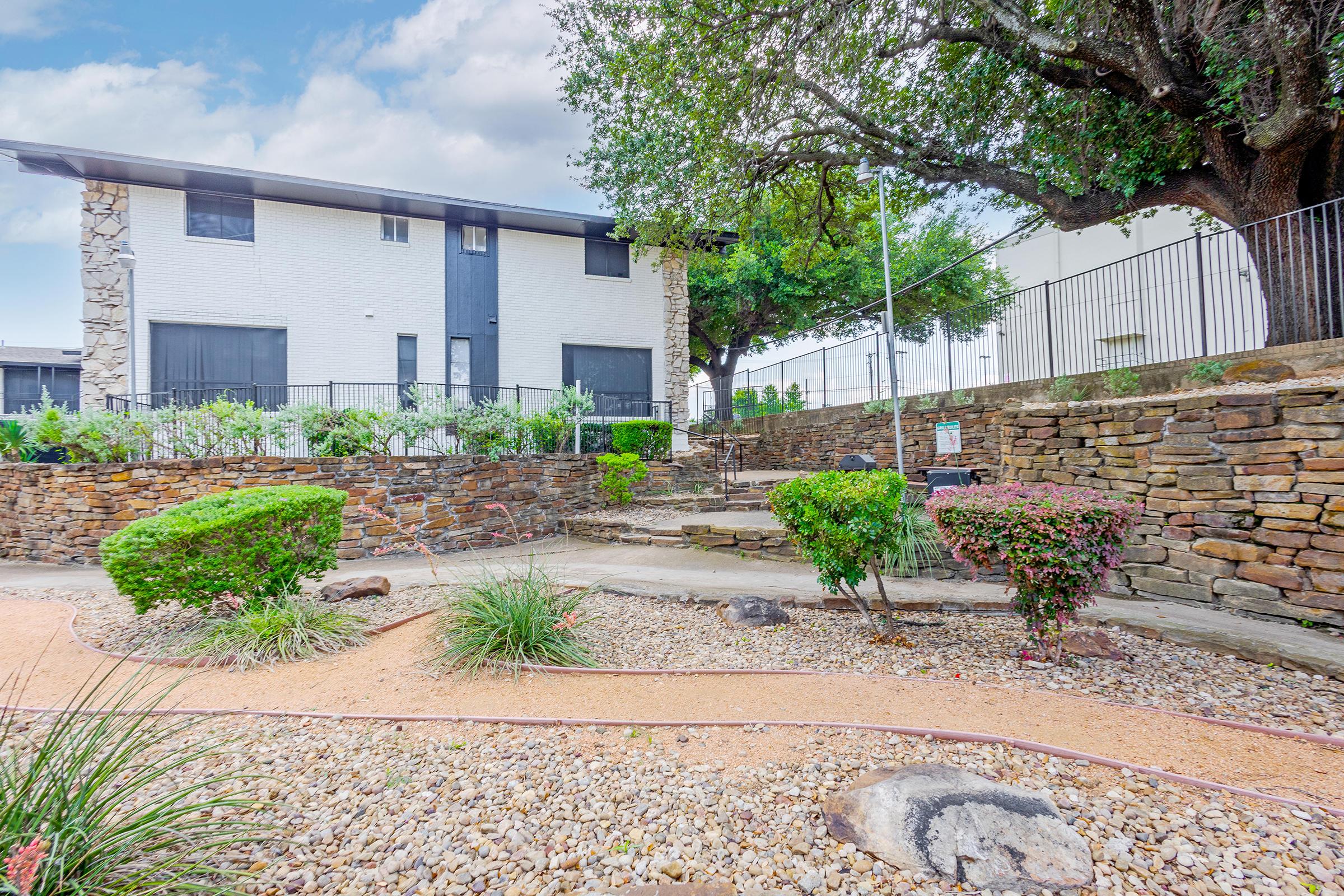 A landscaped area featuring a stone pathway, manicured bushes, and a variety of plants. In the background, there is a multi-story building with a white exterior surrounded by greenery. The scene is bright and airy, with a fence and trees providing a natural setting.