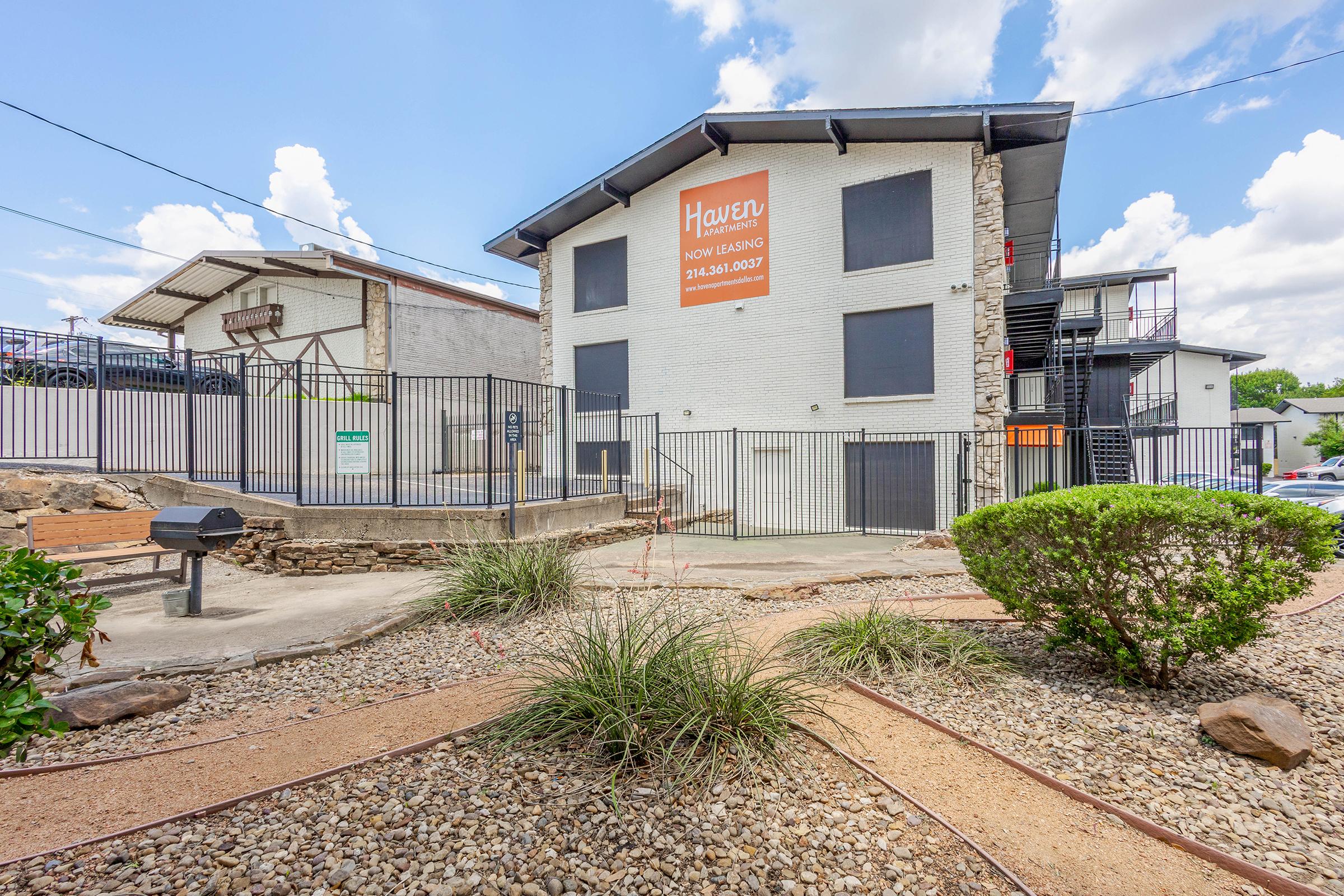 A well-maintained apartment complex featuring a stone exterior, surrounded by landscaped areas with gravel and plants. The building has a sign advertising "Now Leasing" and is equipped with a gated entrance. The sky is partly cloudy, adding a bright atmosphere to the scene.