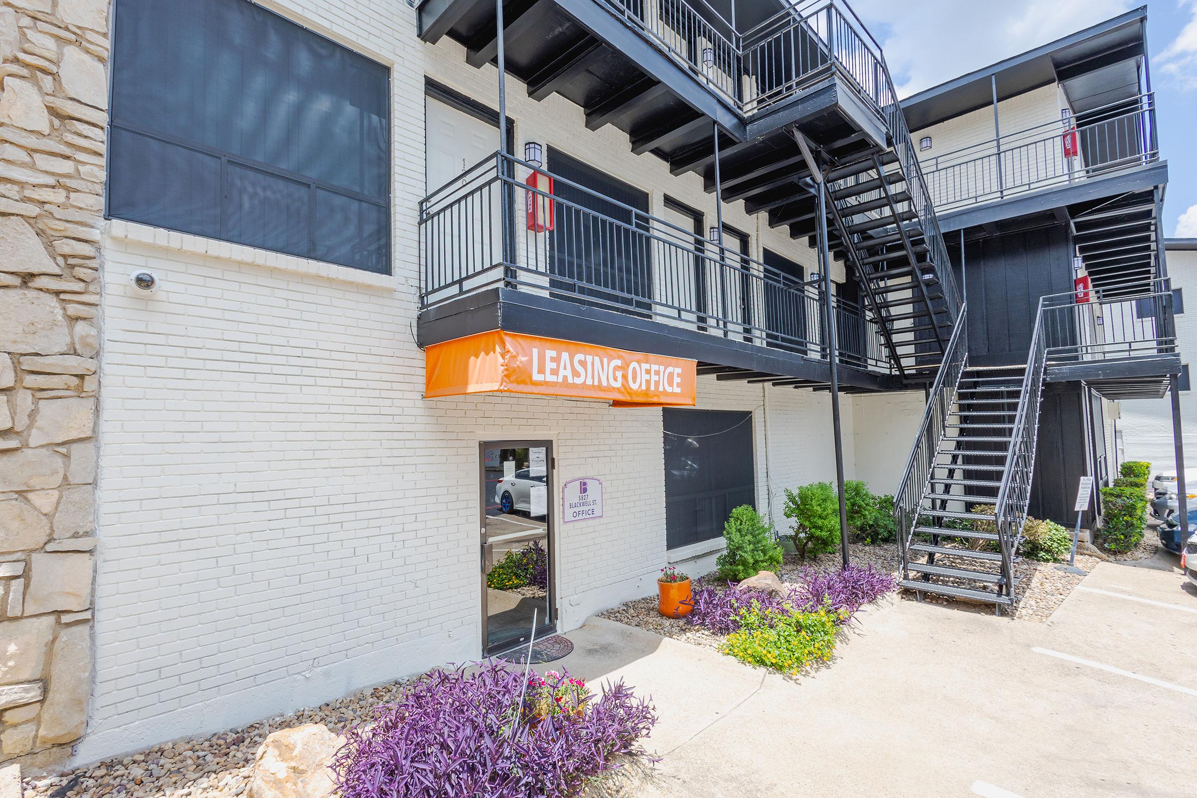 View of a leasing office exterior featuring a bright orange awning with the text "Leasing Office." The building is two stories with black railings and multiple balconies, surrounded by landscaped shrubs and plants. The entrance is visible, along with a sign indicating leasing services.