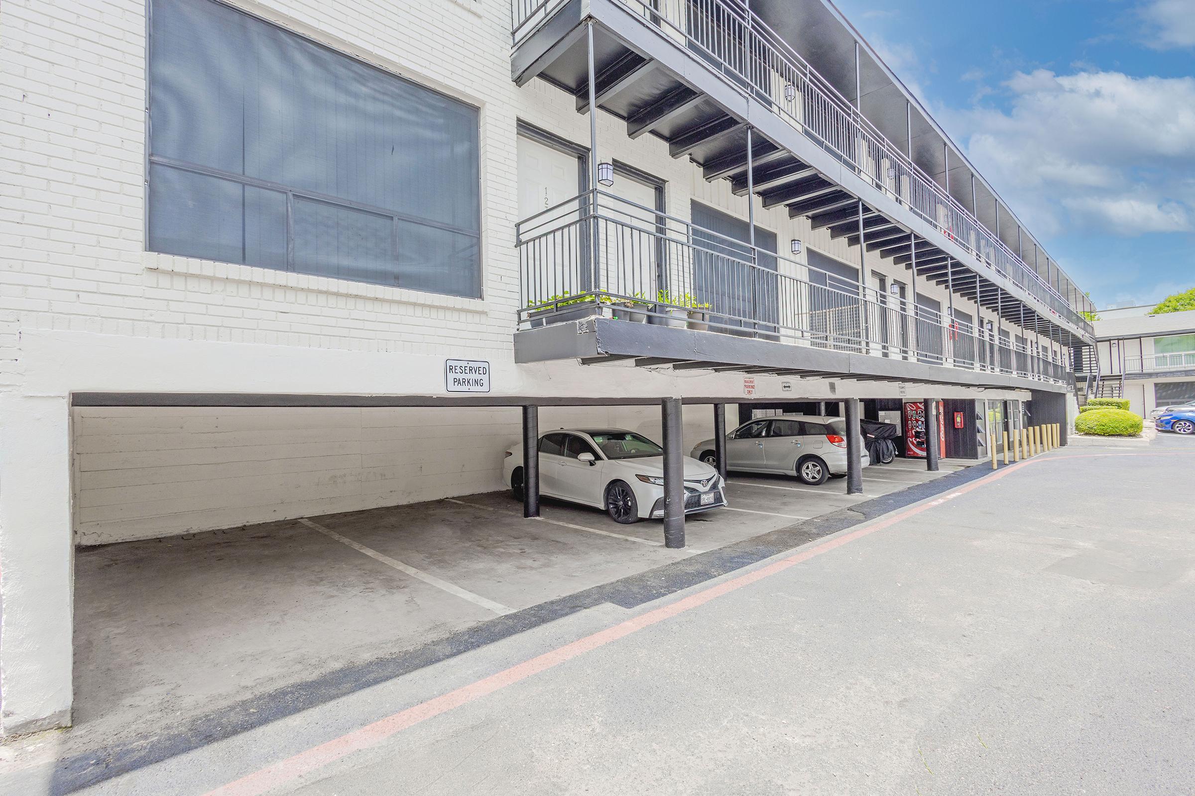 A parking area under a multi-story building, featuring two parked cars and an empty parking space. The building has a white exterior with balconies and a blue sky visible in the background. A sign indicates reserved parking. The area appears clean and organized.