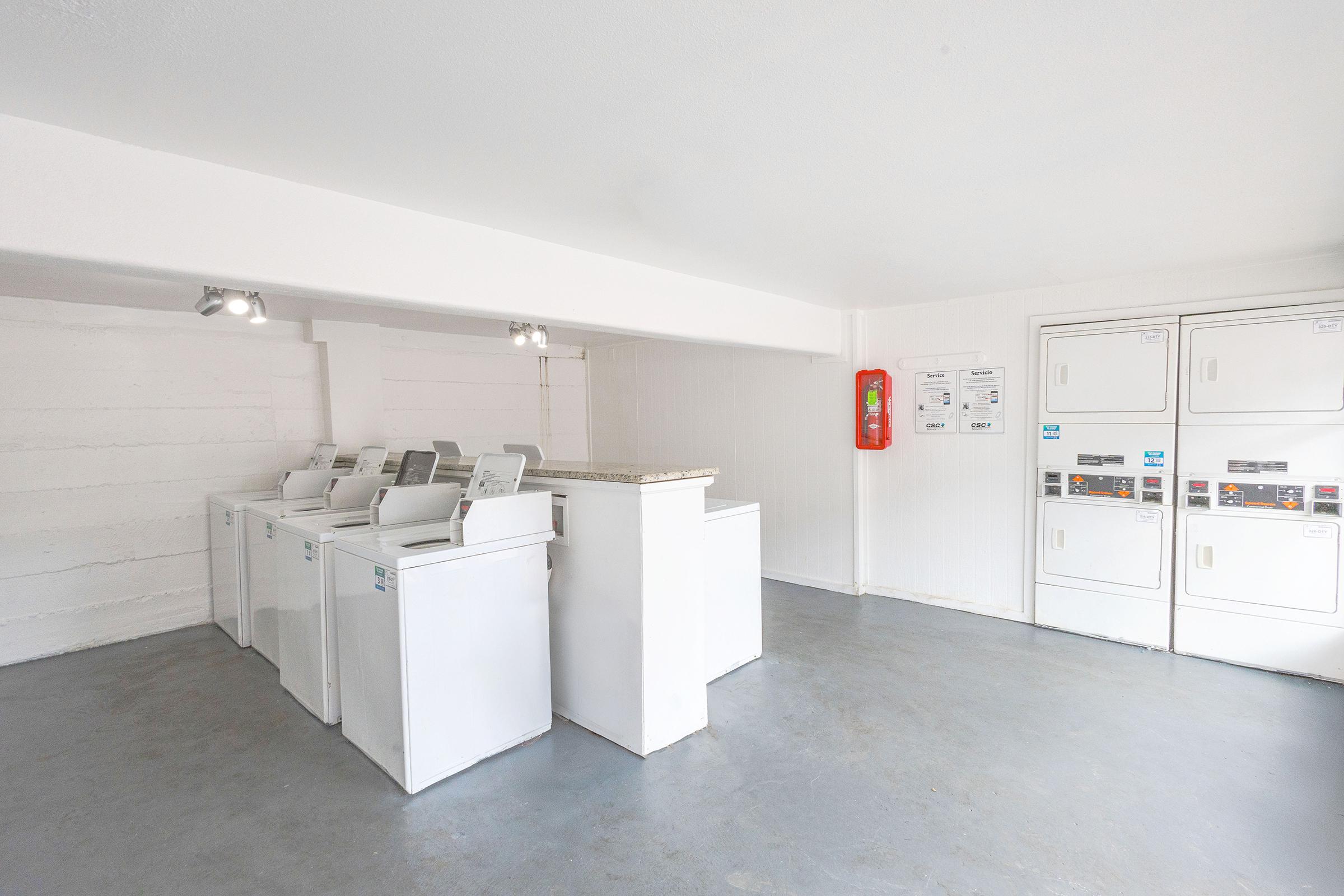 A clean and well-lit laundry room featuring several white washing machines arranged in a row, a countertop area, and multiple dryers. The walls are white, with minimal decor and a fire extinguisher mounted on the wall. The floor is a light gray, creating a modern and tidy atmosphere.