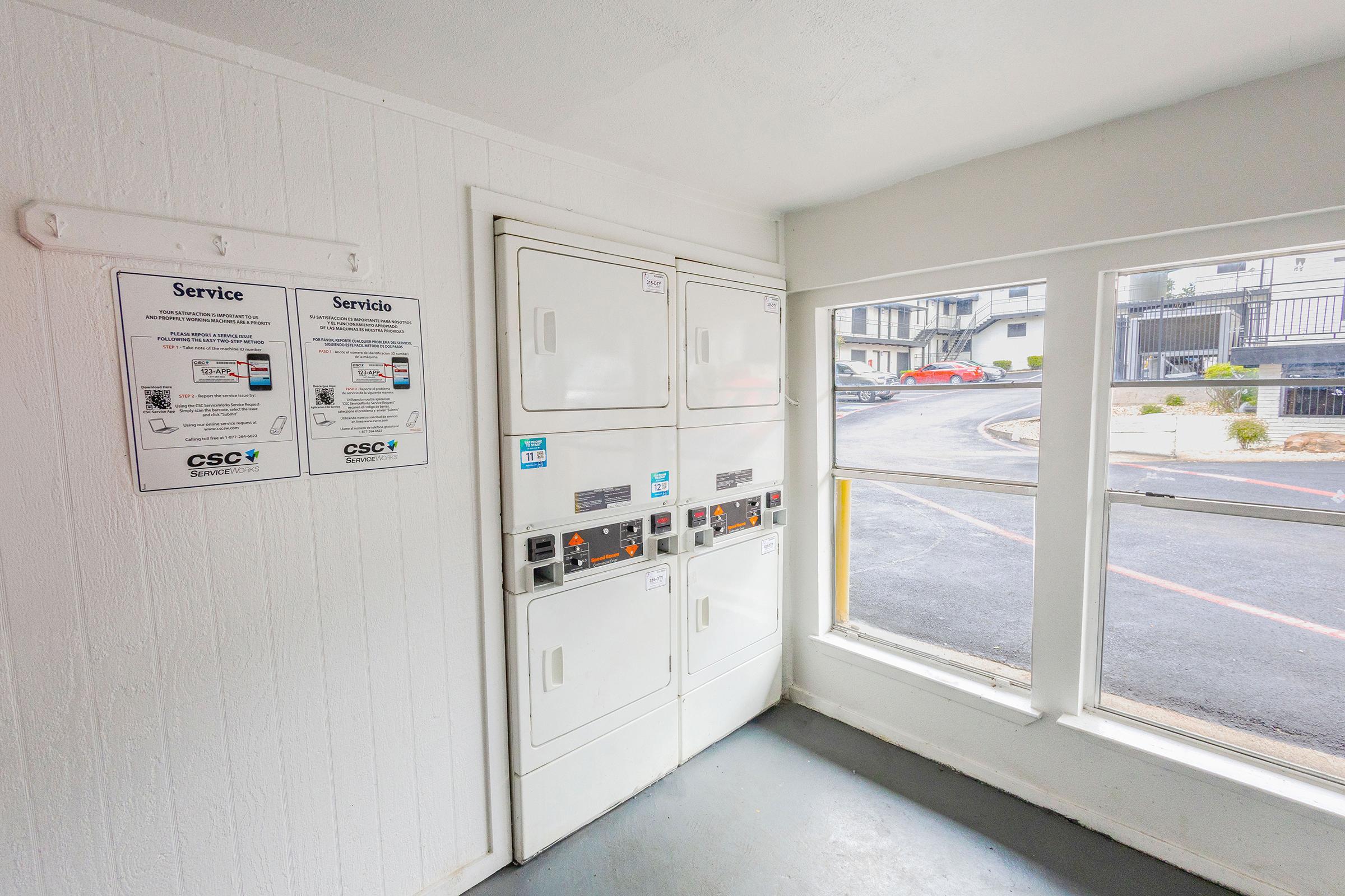 A laundry room featuring stacked washer and dryer units against a white wall. To the left, there are service notices in English and Spanish. Large windows allow natural light, offering a view of the exterior path and landscaping outside. The floor is gray and the overall space is neat and tidy.