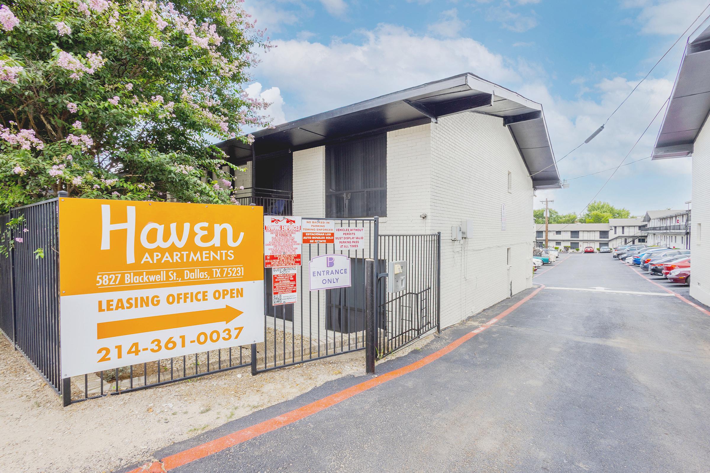 Entrance to Haven Apartments, located at 5827 Blackwell St, Dallas, TX. A visible leasing office sign indicates that it's open. The area features gated access, with parking visible in the background, surrounded by greenery and blooming flowers nearby.