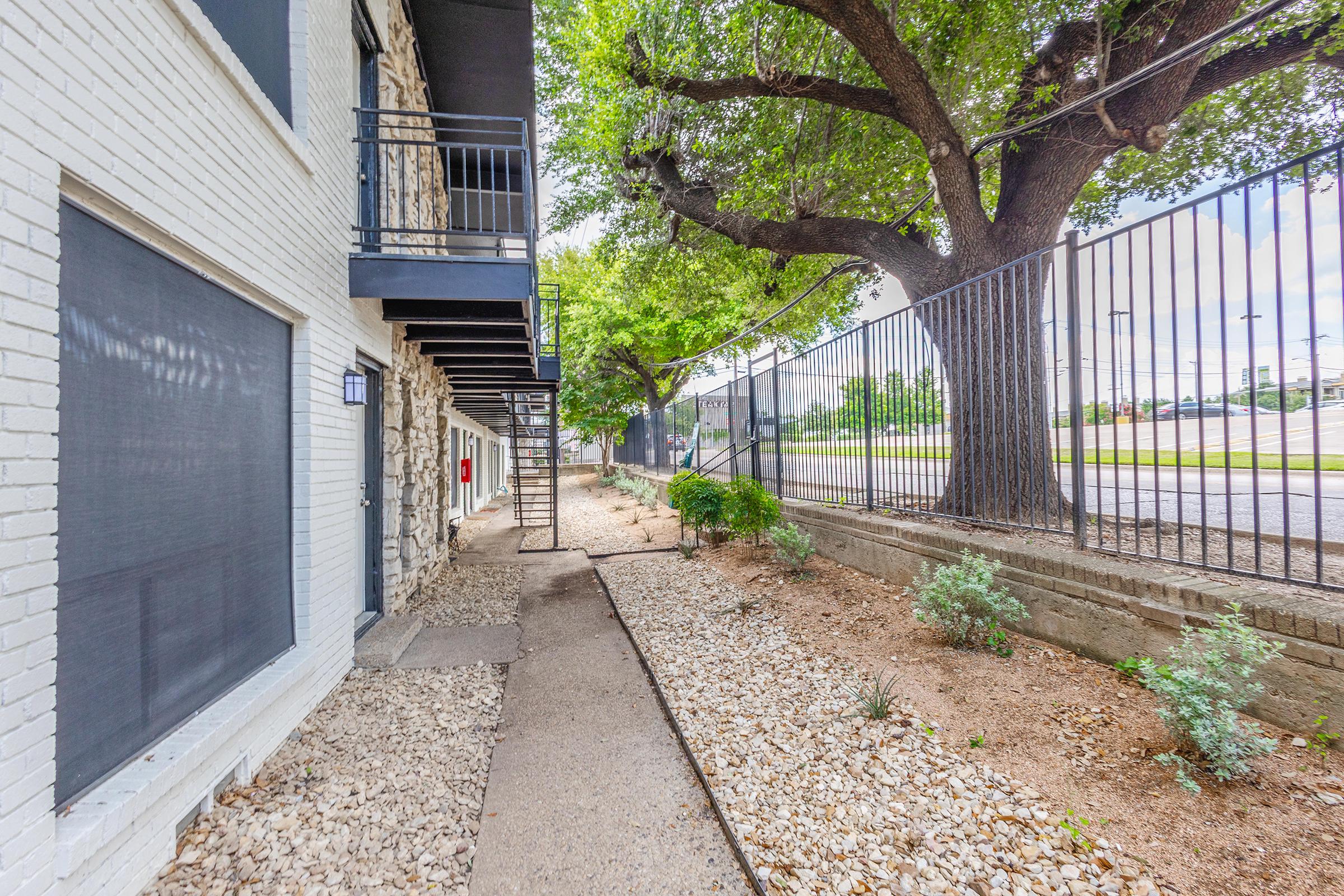 Pathway lined with gravel and small plants alongside a building. There are trees providing shade, and a black metal fence on one side. The scene is bright with a clear sky in the background, suggesting a well-maintained outdoor space near an urban area.