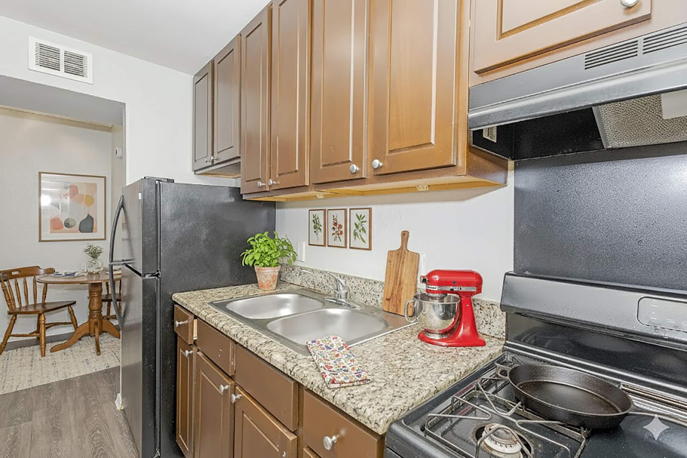 A modern kitchen featuring wooden cabinets, a double sink, a red stand mixer, and a stove with a frying pan. A small potted plant sits on the counter next to a cutting board. In the background, a dining area with a wooden table and chairs is visible.