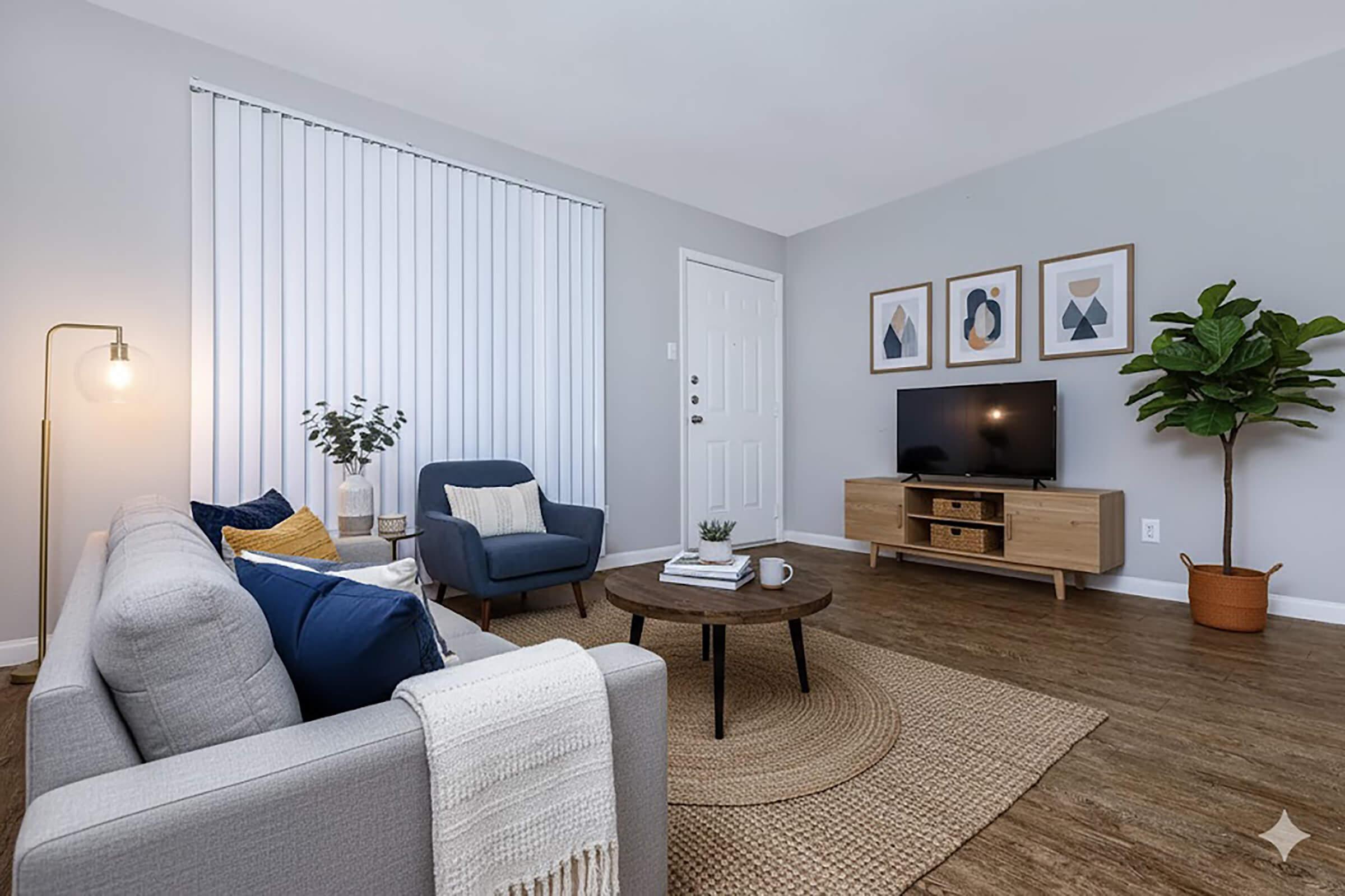 A cozy living room featuring a light gray sofa with blue and yellow cushions, a round coffee table, and a modern TV unit with a television. There are three framed geometric artworks on the wall, a plant in a pot, and a window with vertical blinds, creating a warm and inviting atmosphere.
