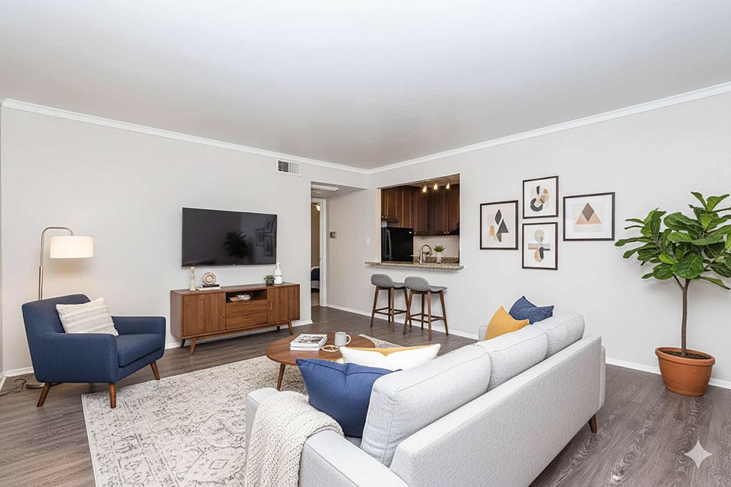 A modern living room featuring a light gray couch with blue and yellow cushions, a wooden coffee table, and a stylish armchair. A large television is mounted on the wall across from the seating area. The space is decorated with framed artwork and includes a potted plant, creating a cozy and inviting atmosphere.