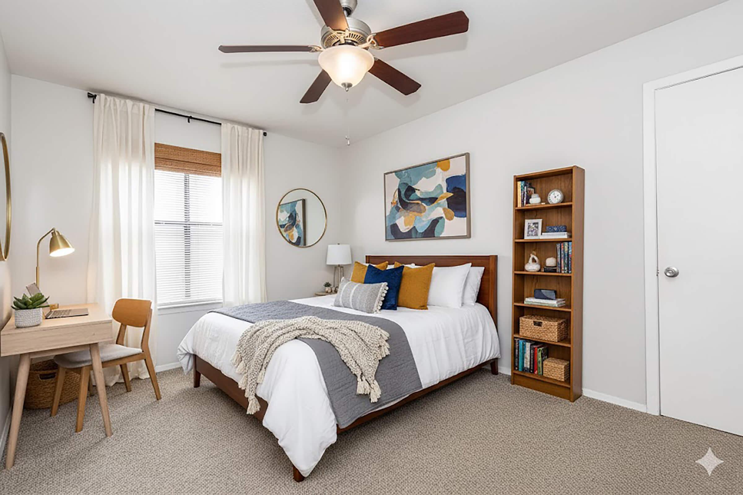 A cozy bedroom featuring a wooden bed with grey and white bedding, accented with colorful pillows. A small desk with a chair is positioned next to the window, and a ceiling fan is overhead. A bookshelf displays decorative items, and a piece of abstract art hangs on the wall, complemented by soft natural light.