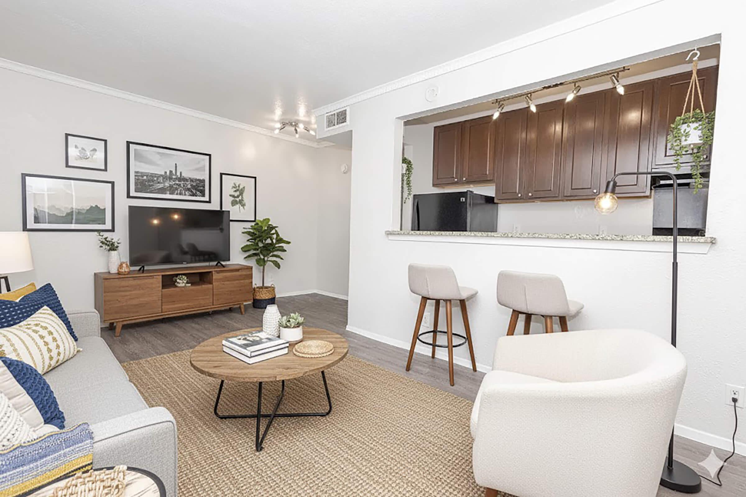 A modern living room featuring a light gray sofa with decorative pillows, a round coffee table on a woven rug, and a TV on a wooden media console. In the background, there's a kitchen bar with two stools, dark cabinetry, and plants, creating a welcoming and stylish atmosphere.