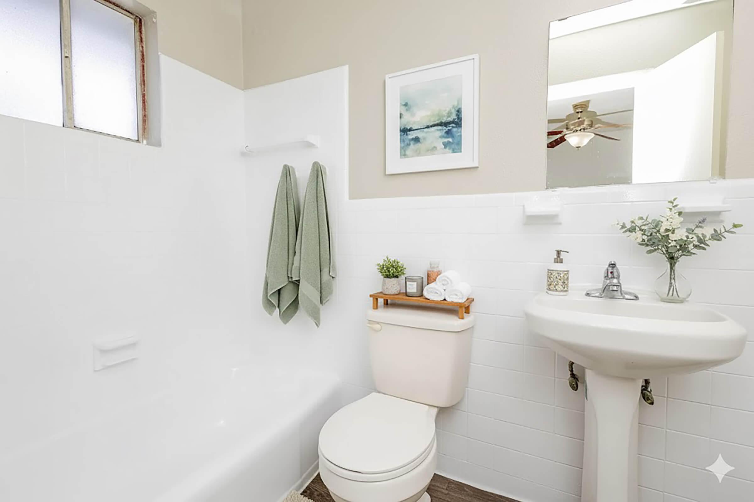 A clean, modern bathroom featuring a bathtub with a white surround, a white toilet, and a pedestal sink. There are two green towels hanging on a rack, and a small shelf with neatly arranged toiletries. A mirror reflects the room, and a light fixture is visible on the ceiling.