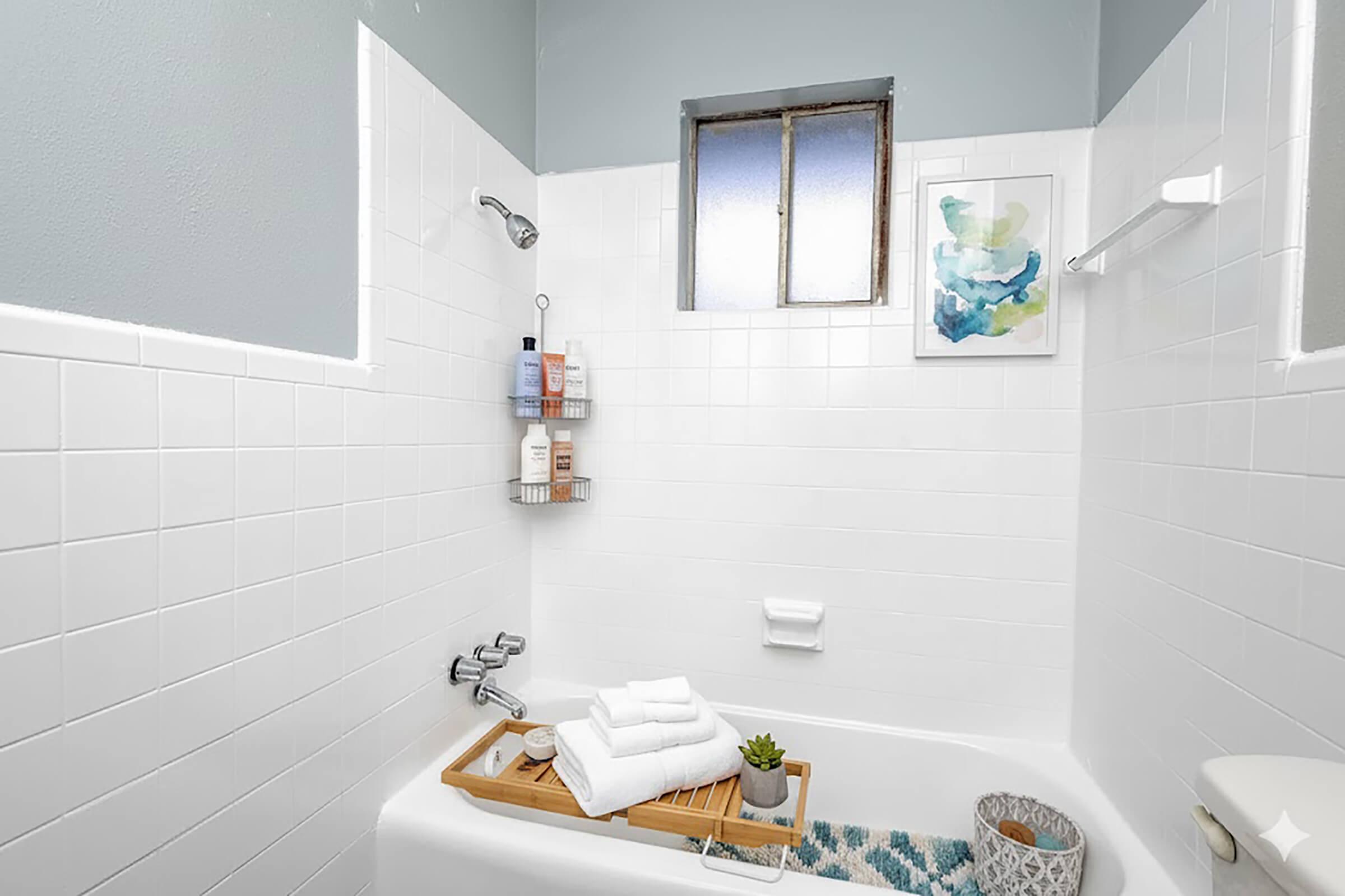 A neatly arranged bathroom featuring a white tiled shower area, a showerhead, and a small window. There are neatly stacked white towels on a wooden tray, a small plant, and a decorative rug. A wall-mounted basket holds toiletries, and a piece of art is hung above the bath area.