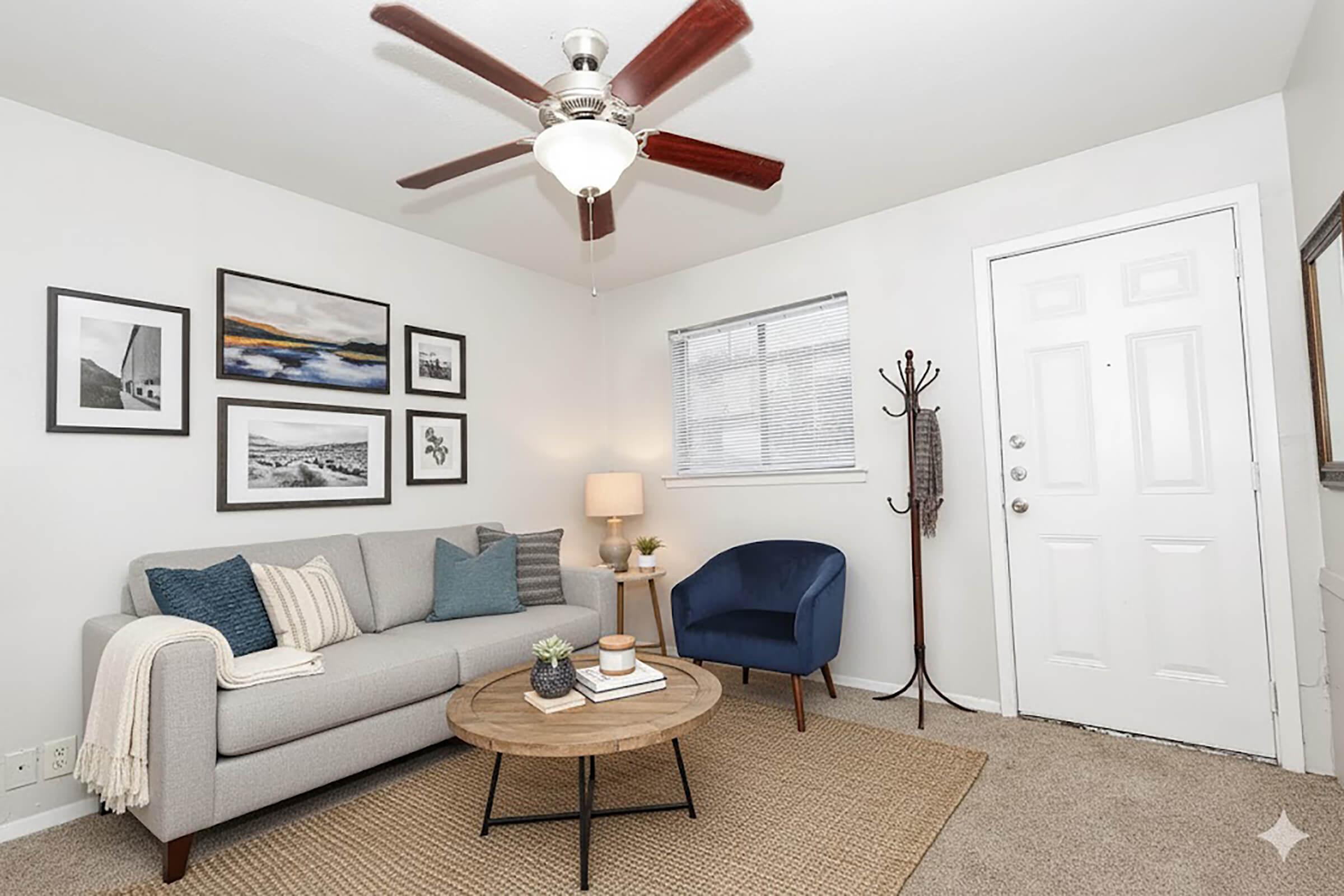 Cozy living room featuring a light gray sofa with decorative pillows, a round coffee table, and an inviting blue chair. The walls are adorned with framed black-and-white photos and a landscape print. A ceiling fan provides a modern touch, and a door leads outside. Natural light enters through a window, enhancing the warm atmosphere.