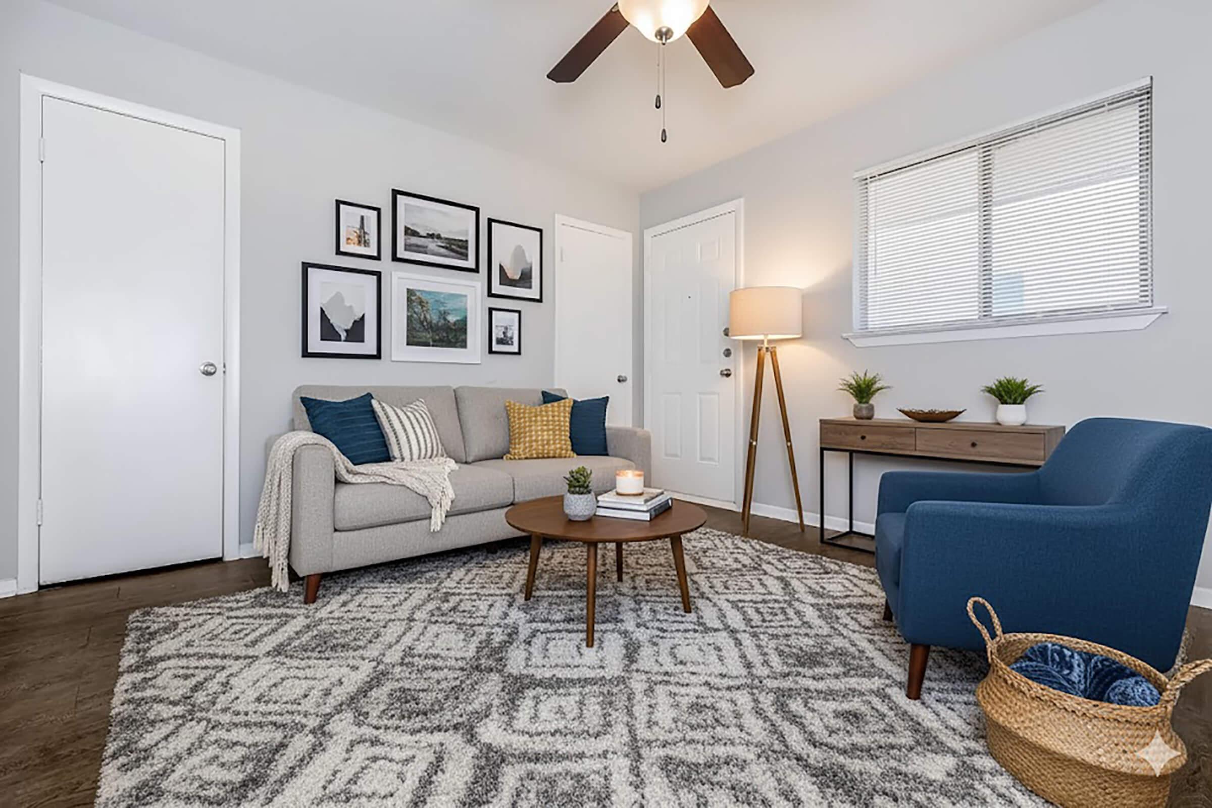 A cozy living room featuring a gray sofa with blue and yellow pillows, a round coffee table with a candle, and a textured area rug. There are framed pictures on the wall and a small lamp beside a wooden side table. A door and a window provide natural light to the space.