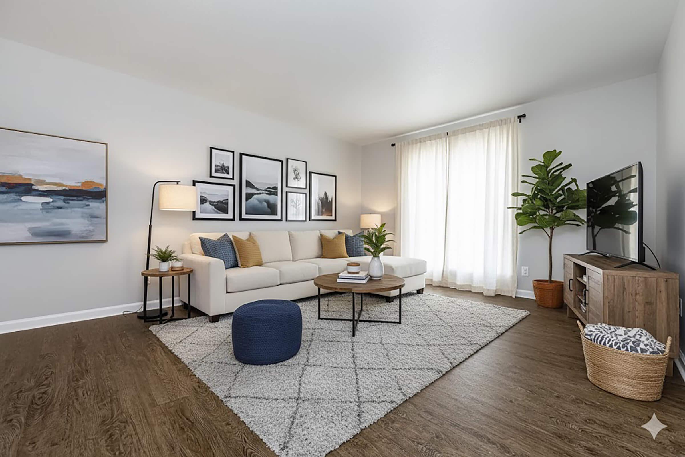 A modern living room featuring a comfortable white sectional sofa with patterned throw pillows, a wooden coffee table, and a large TV. Natural light streams in through sheer curtains, illuminating the space, which includes a potted plant, decorative wall art, and a cozy rug.