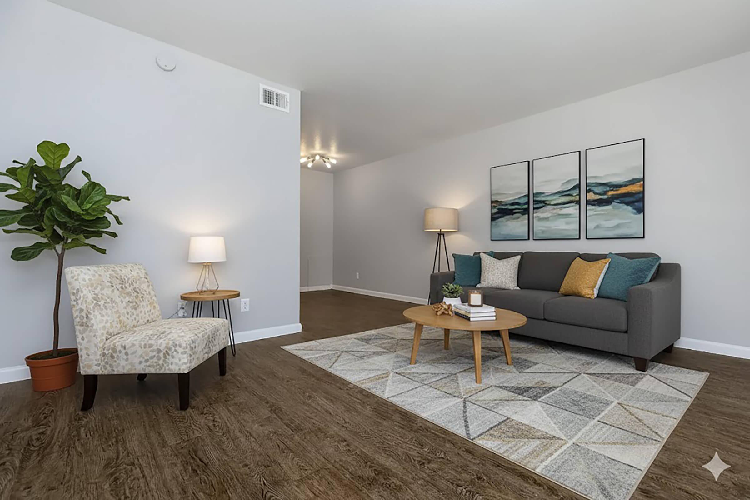 A modern living room featuring a gray sofa with decorative pillows, a light-colored armchair, and a round coffee table. The space includes a patterned area rug and a potted plant. Wall art consisting of three abstract paintings adds color, while lamps provide soft lighting. The floor is a warm wood tone.