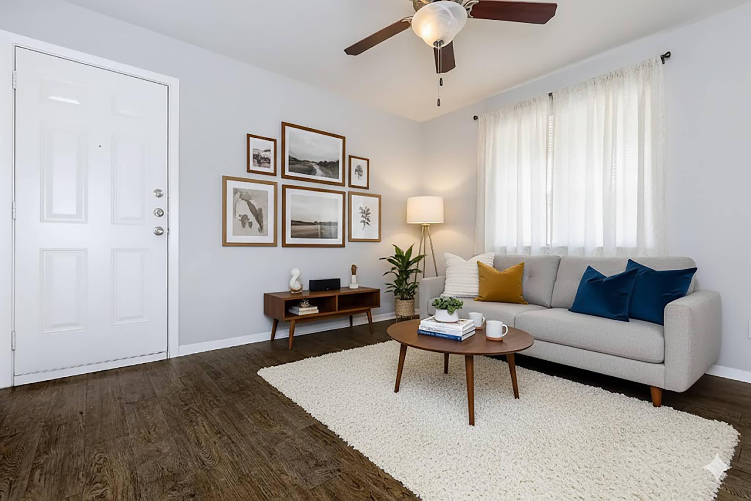 A cozy living room featuring a light gray sofa adorned with blue and gold accent pillows. A wooden coffee table sits on a white plush rug, with a small plant and a coffee cup on it. The walls are decorated with framed art, and a floor lamp provides warm lighting. A door and window allow natural light to enter.