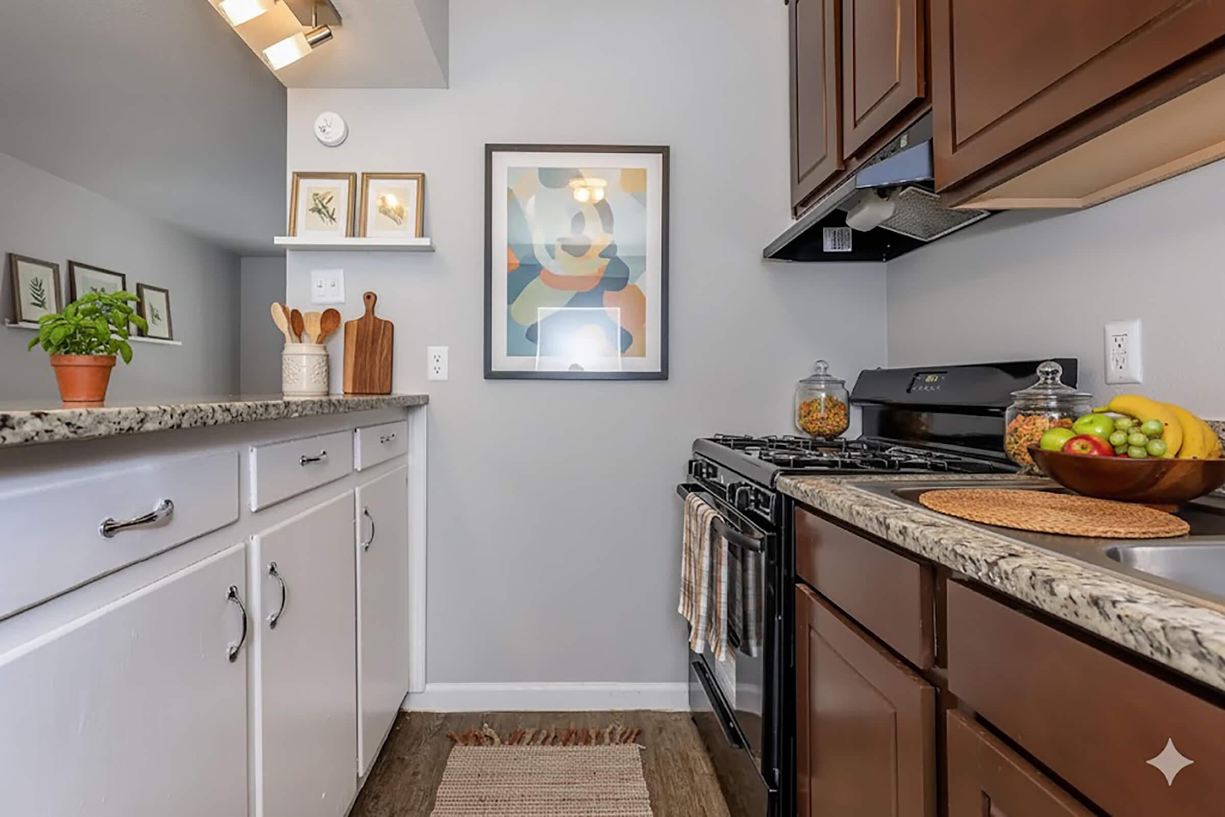 Modern kitchen featuring dark wood cabinets and granite countertops. A sleek black stove and a round woven placemat with colorful fruits on one side. On the opposite side, white cabinets with wooden utensils displayed in a jar and framed artwork on the wall. Natural light enhances the warm, inviting atmosphere.