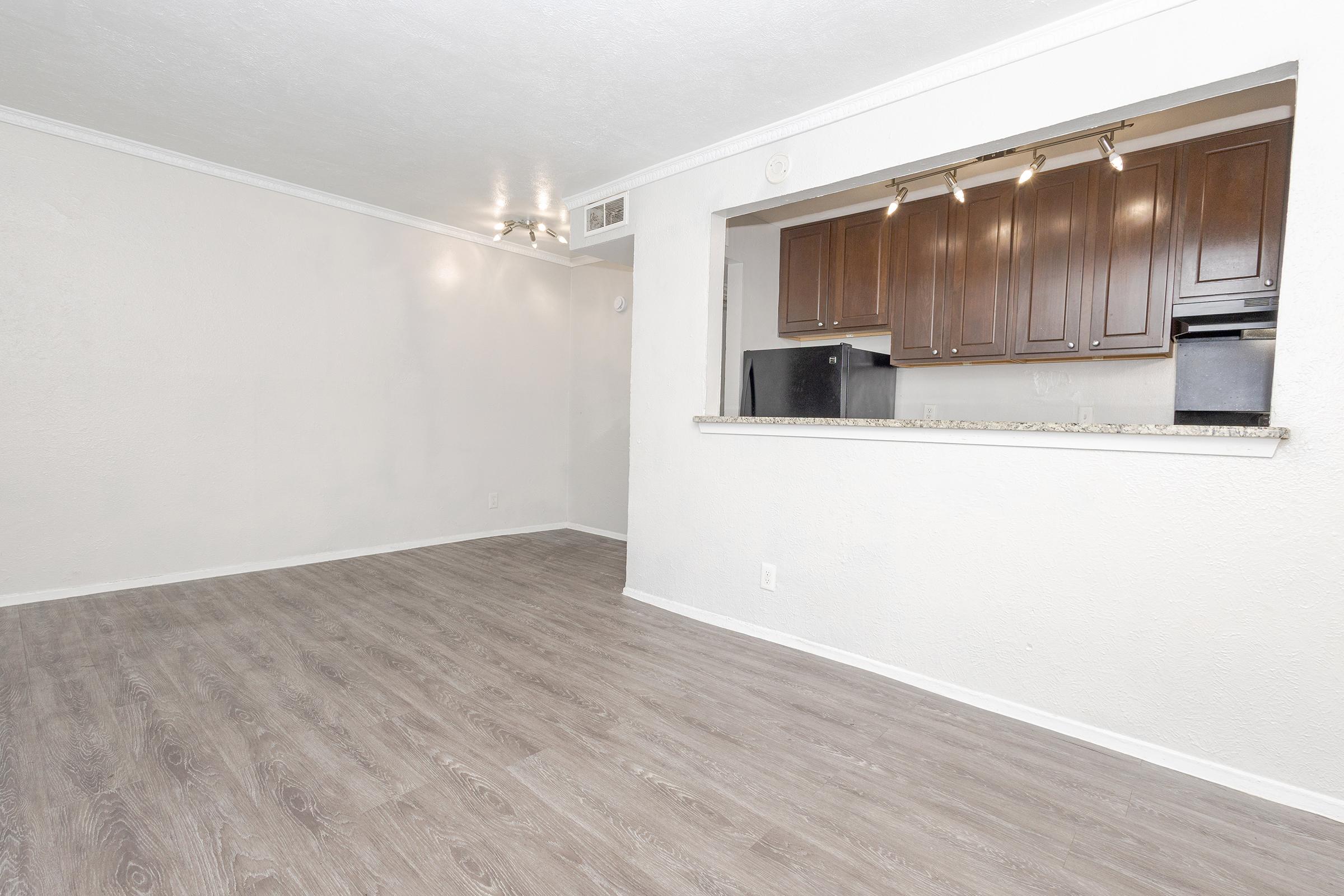A vacant room featuring light-colored walls and wood laminate flooring. An open kitchen area with dark wooden cabinets is visible in the background. The space is well-lit, creating a bright and airy atmosphere.