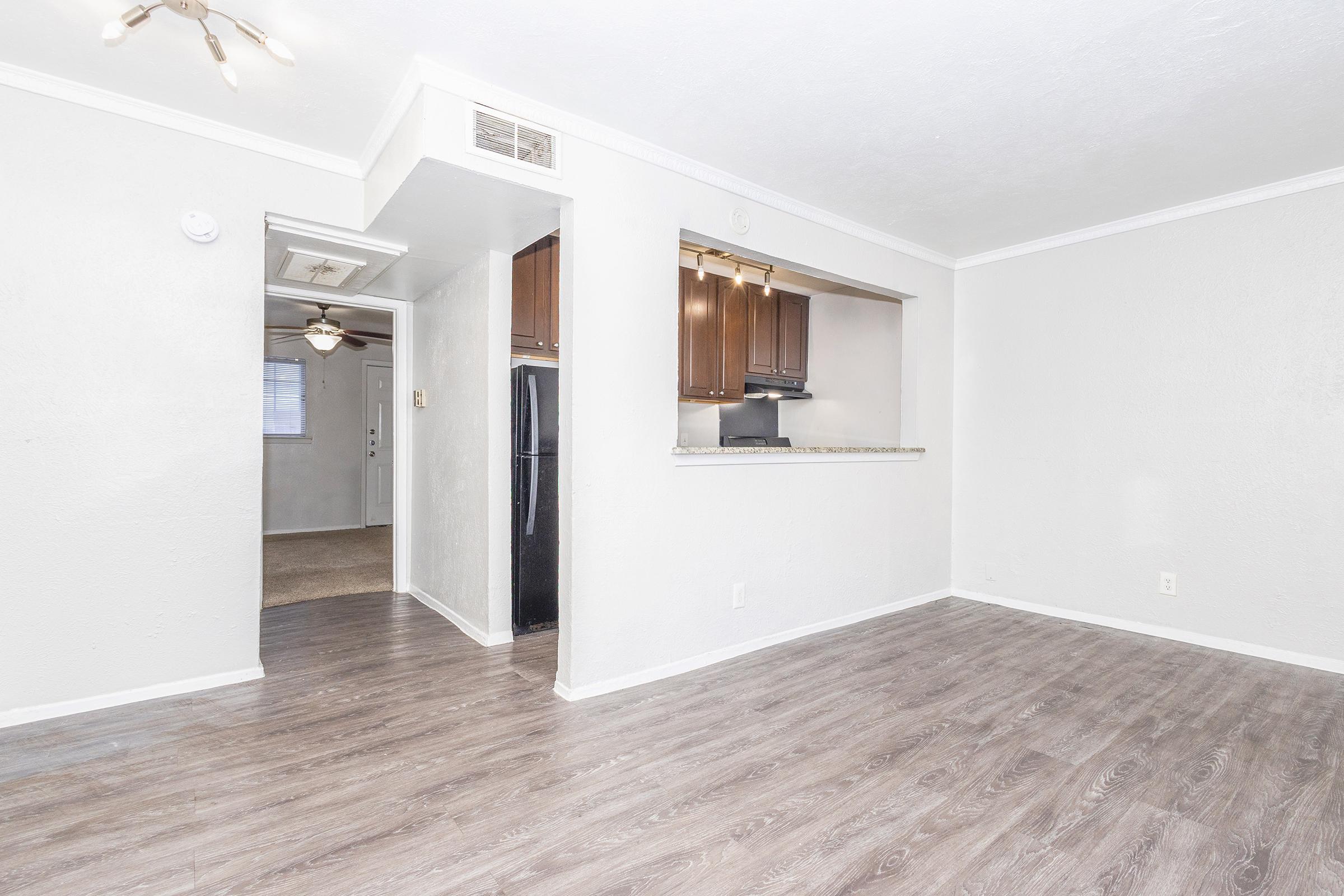 A spacious living area with light gray walls and laminate flooring, featuring an open layout that connects to a kitchen with dark wood cabinets. A doorway leads to another room, and there is a ceiling fan overhead. Natural light filters in, creating a bright and inviting atmosphere.