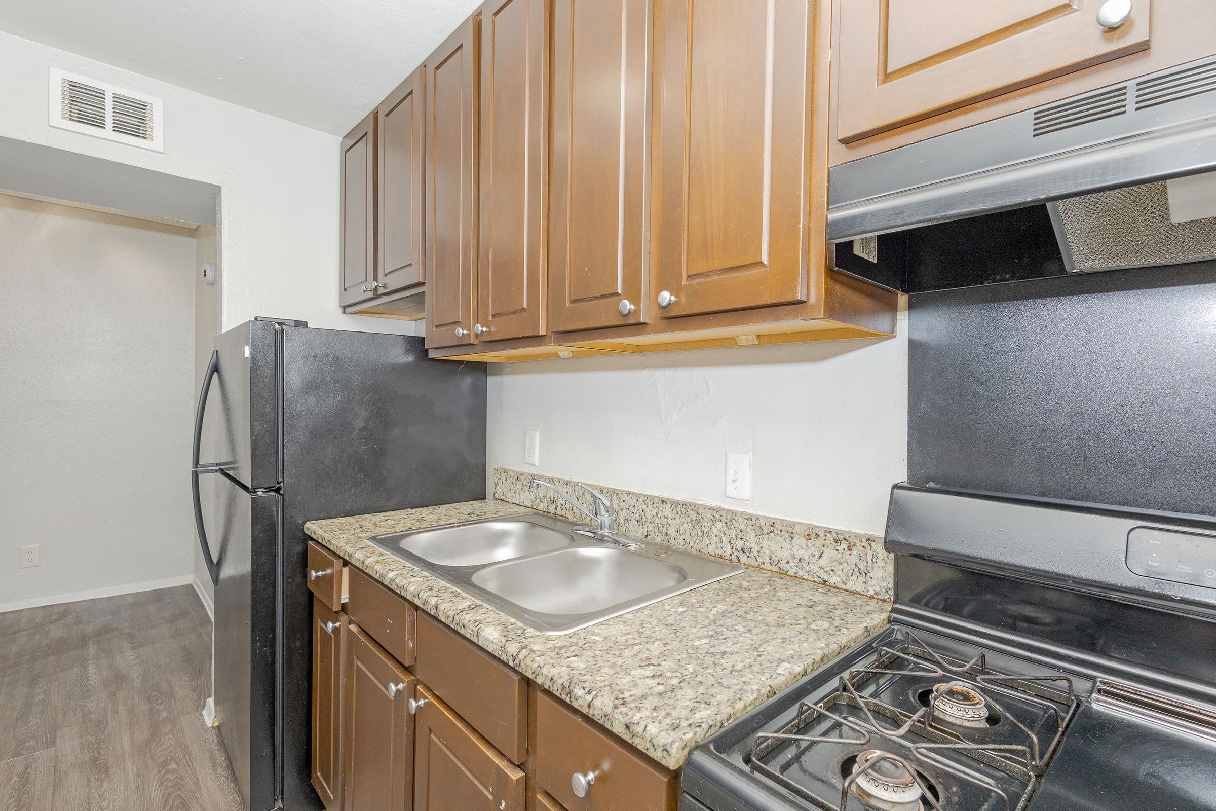 A small kitchen featuring dark wooden cabinets, a double sink, a granite countertop, and a black refrigerator. The kitchen also includes a black stove with an oven and a hood above. The flooring is a dark, wood-like material, and there's an open doorway leading to another room.
