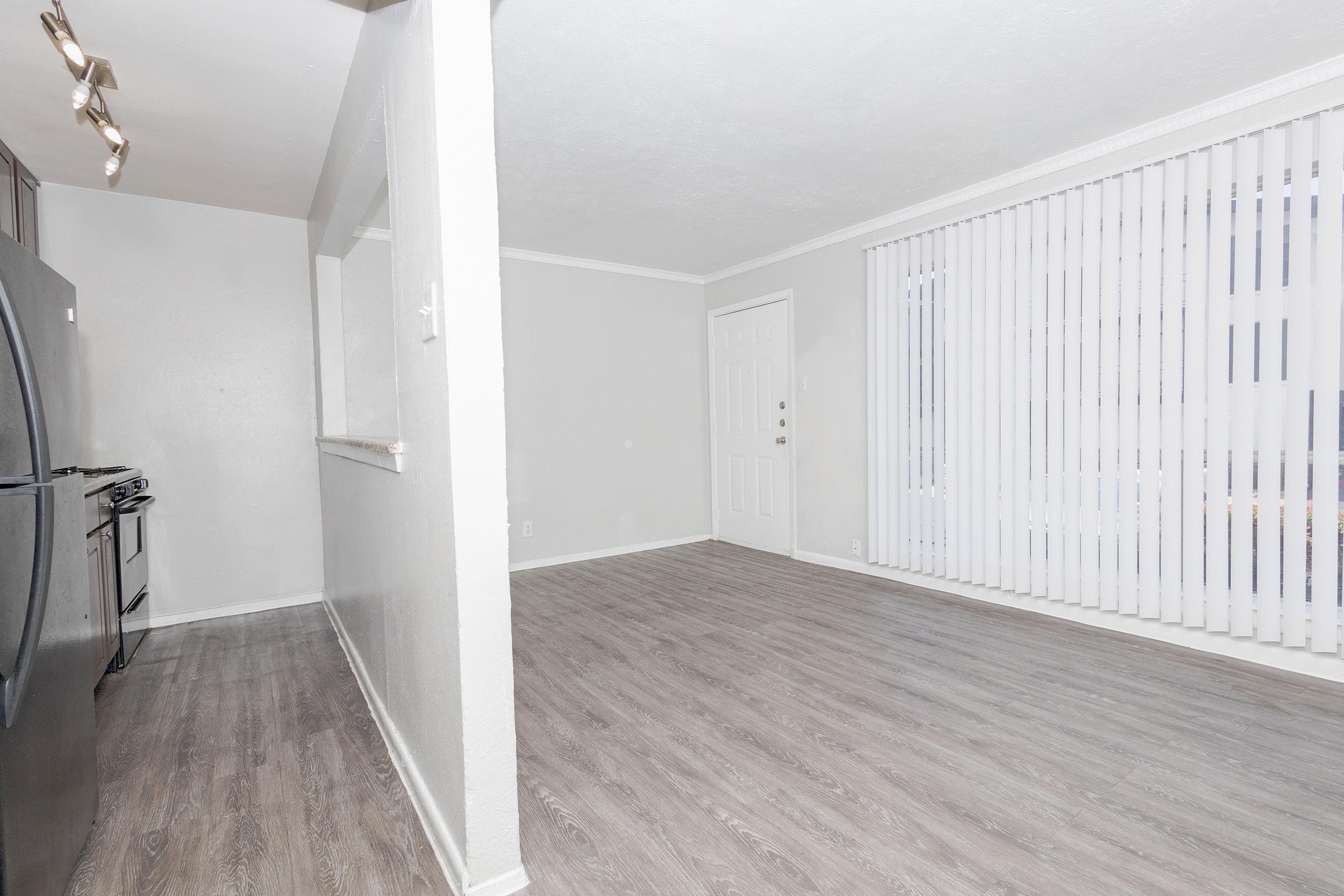 An empty living room space with light gray walls and light wood flooring. There is a large window with vertical blinds allowing natural light in, and a doorway leading to another room. A black refrigerator and stove are visible in the kitchen area to the left. The space appears clean and minimalistic.