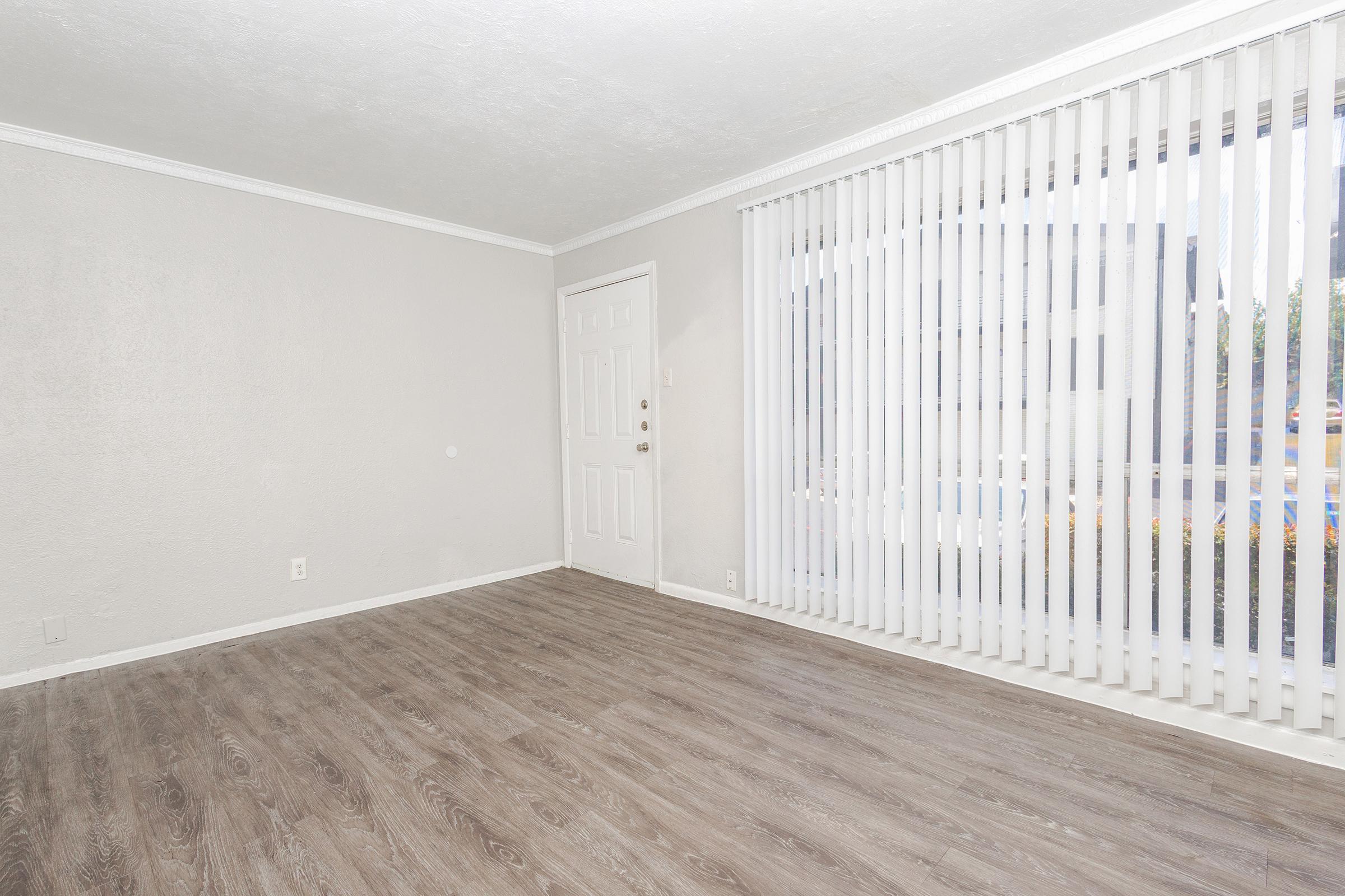 An empty room with light-colored walls and a wood-like floor. A large window with vertical blinds allows natural light to enter. The door is located on the left side, while the rest of the space remains bare, creating a minimalist atmosphere.