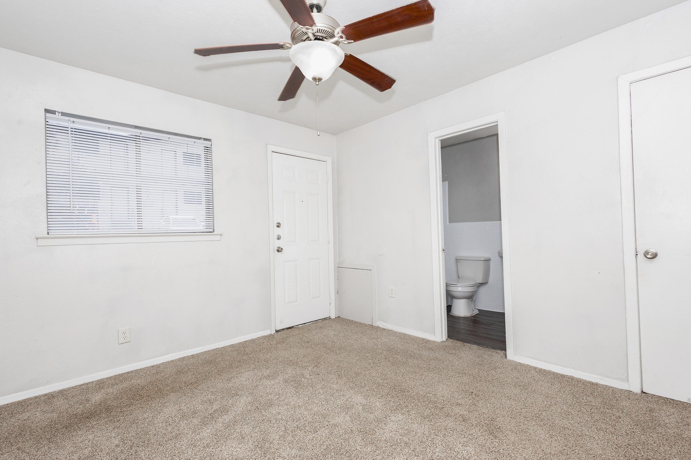 A spacious room featuring beige carpet, a ceiling fan, and a window with blinds. There is a white door leading outside and a visible bathroom door to the right. The walls are painted white, creating a bright and airy atmosphere.