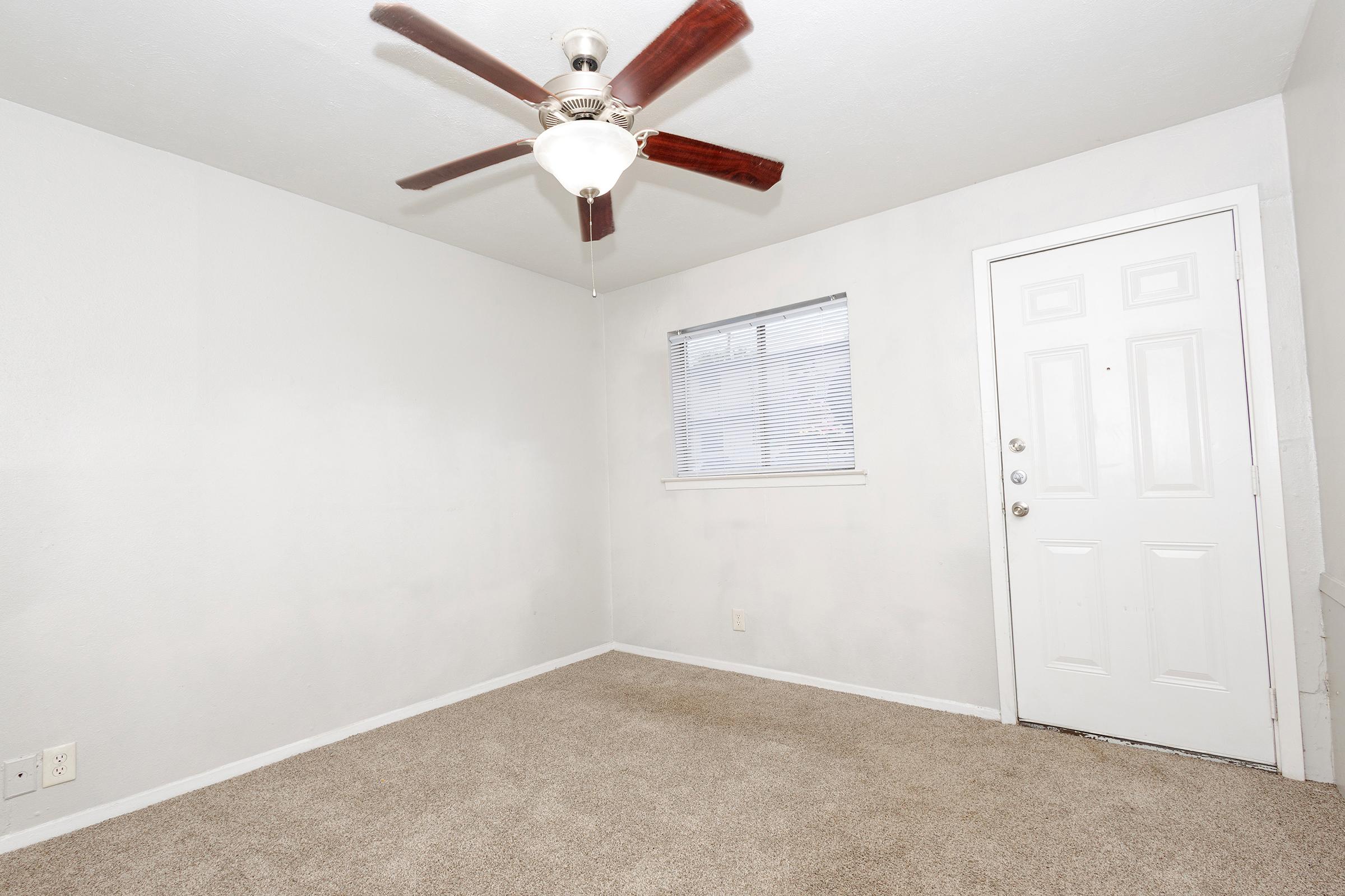Empty room featuring light beige carpet, a ceiling fan with a light fixture, a window with blinds, and a white door. The walls are painted a light color, and there is no furniture present, creating a minimalist and open atmosphere.